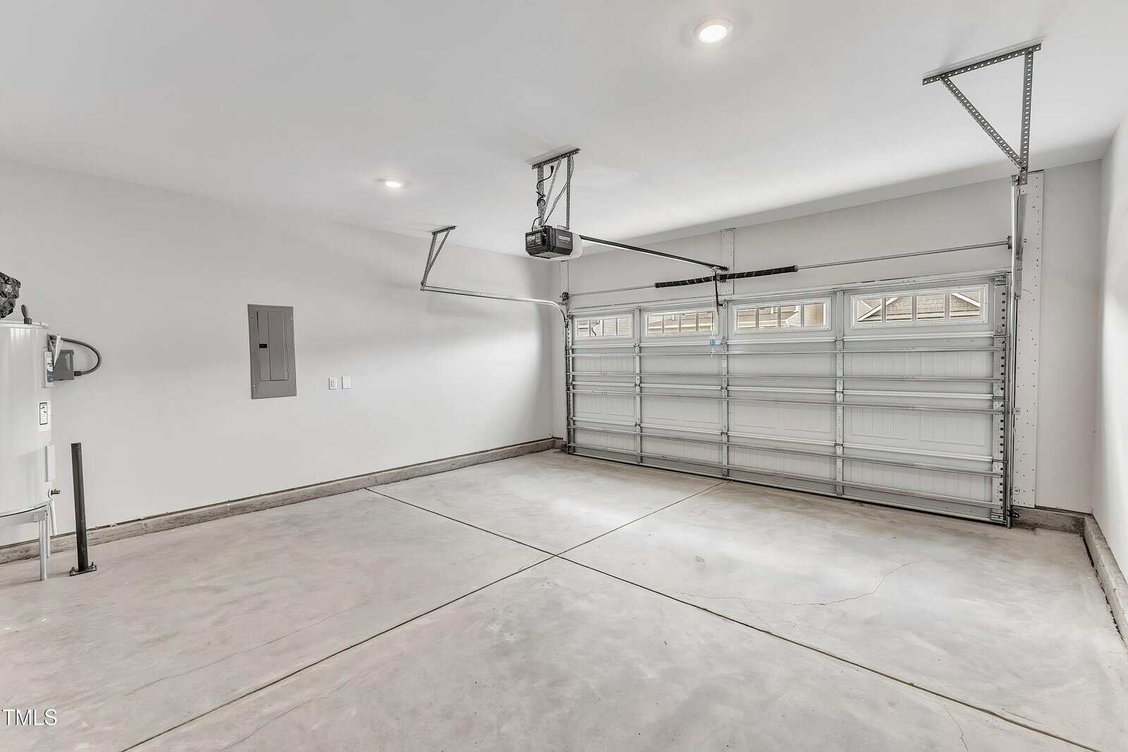 A clean, empty two-car garage with a closed sectional garage door, ceiling-mounted opener, bright overhead lights, and light gray walls. There is an electrical panel and a water heater on the left wall.