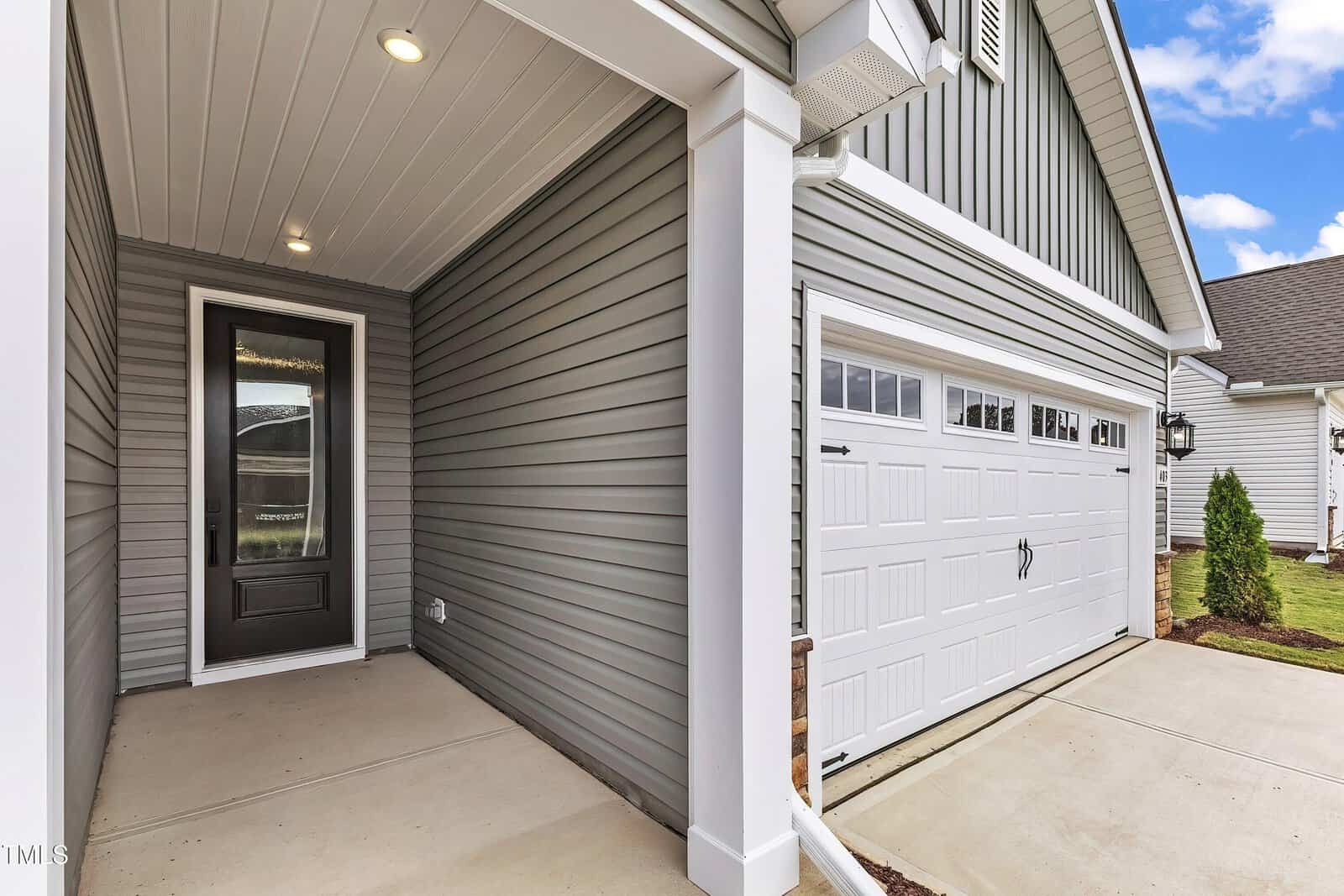 Covered front porch with a black glass-paneled door next to a white two-car garage; gray vinyl siding and concrete driveway; small patch of grass and blue sky visible.