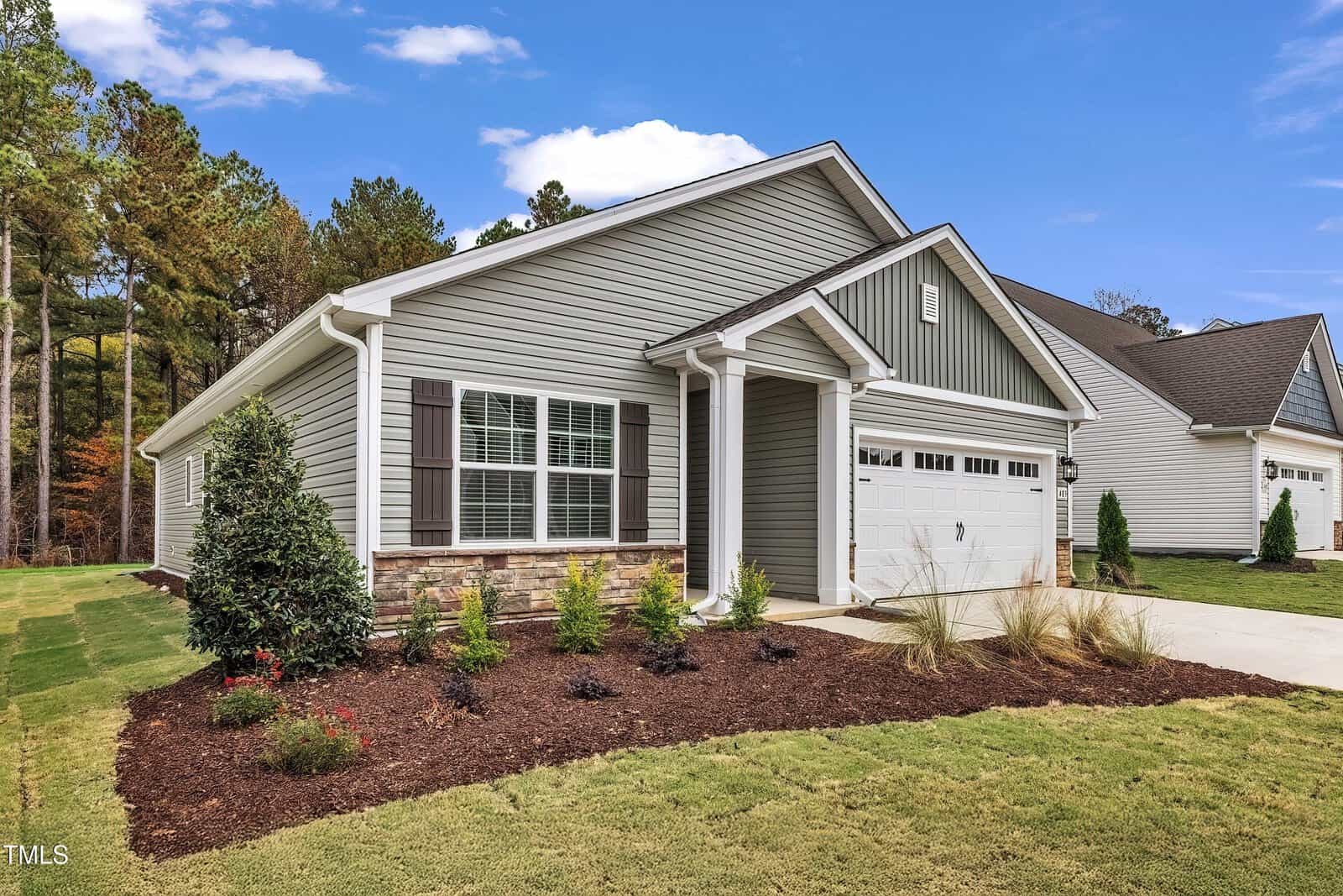 Single-story suburban house with light gray siding, white trim, and a gabled roof. There are brown shutters, a front porch with columns, landscaped garden beds, and a two-car garage. The lawn is neatly maintained.