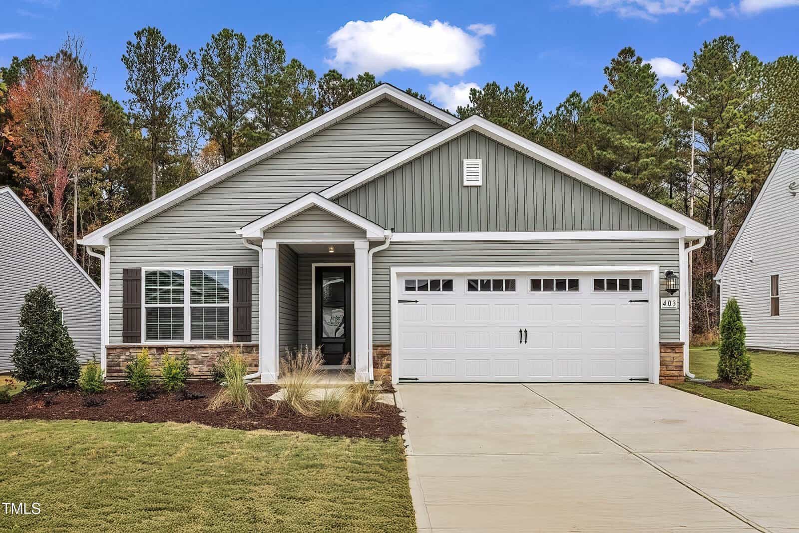 Single-story suburban house with gray siding and white trim, double garage door, covered front porch, manicured lawn, shrubs, and trees in the background under a partly cloudy sky.
