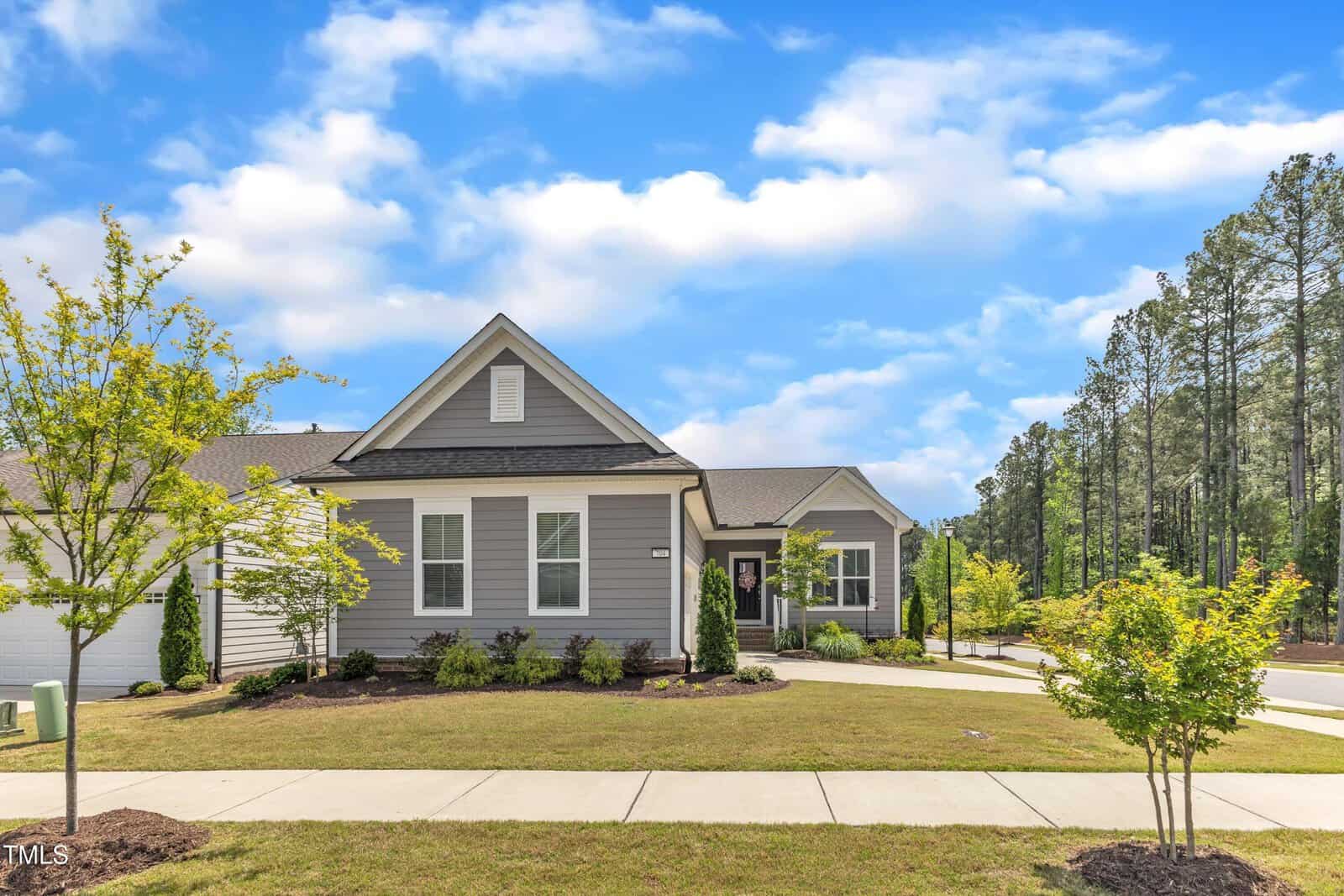 A single-story gray house with white trim, neatly landscaped yard, trees, and a sidewalk in front. The sky is bright blue with some clouds, and tall pine trees are visible to the right.