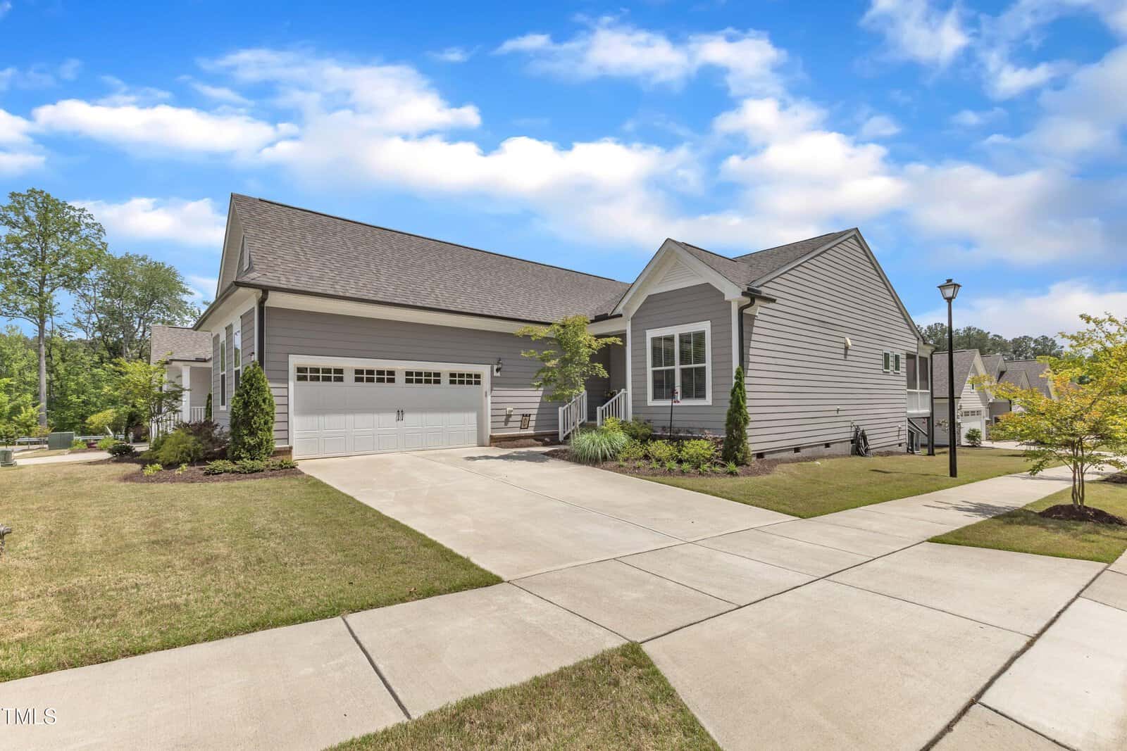 A modern single-story gray house with a two-car garage, sloped roof, and neatly landscaped yard sits on a sunny day with a blue sky and scattered clouds. A wide driveway and sidewalk run alongside the home.