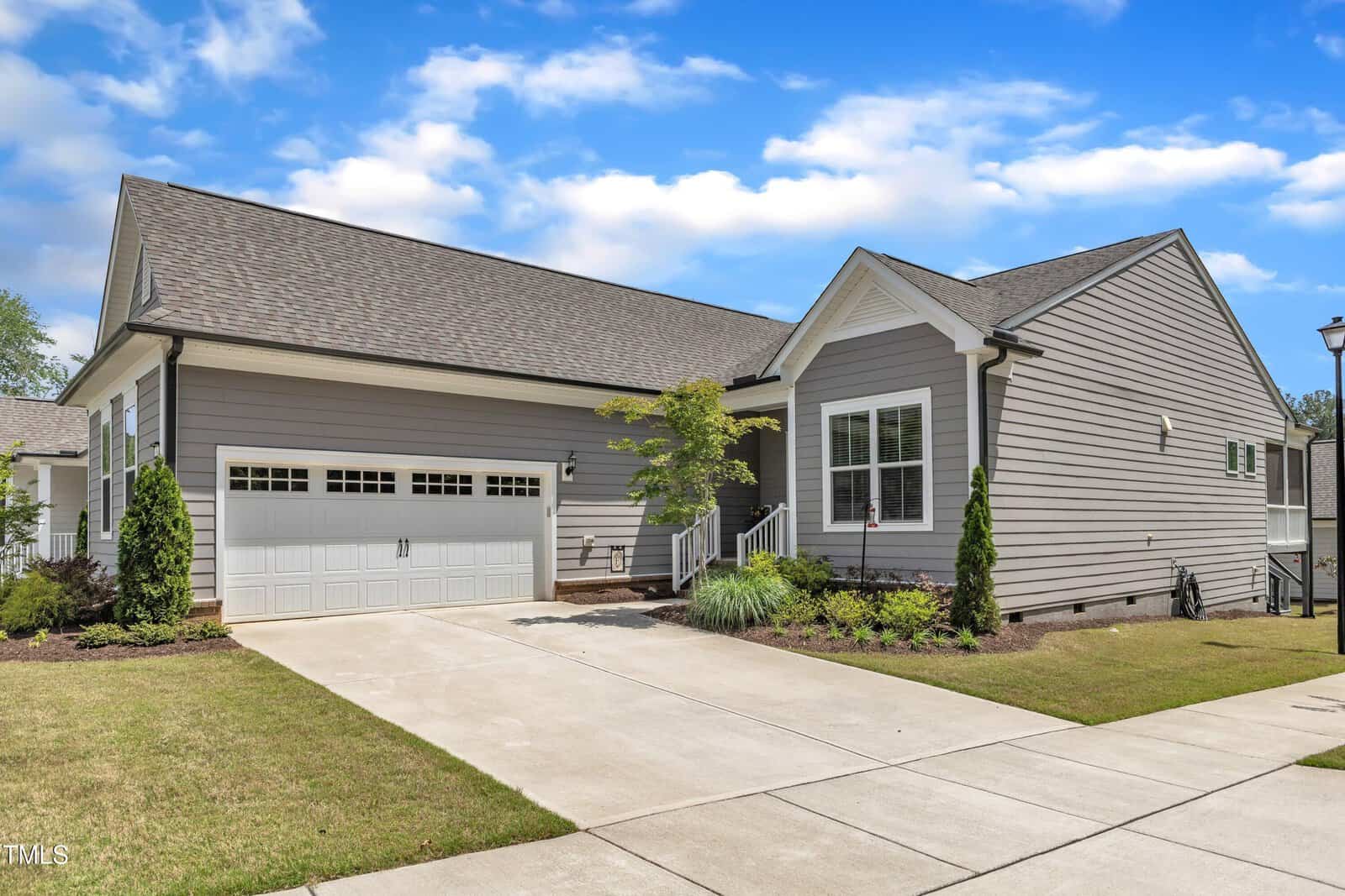 A modern single-story house with gray siding, white trim, and a double garage. The driveway and front lawn are neatly maintained, and there are small shrubs and trees near the entrance on a sunny day.
