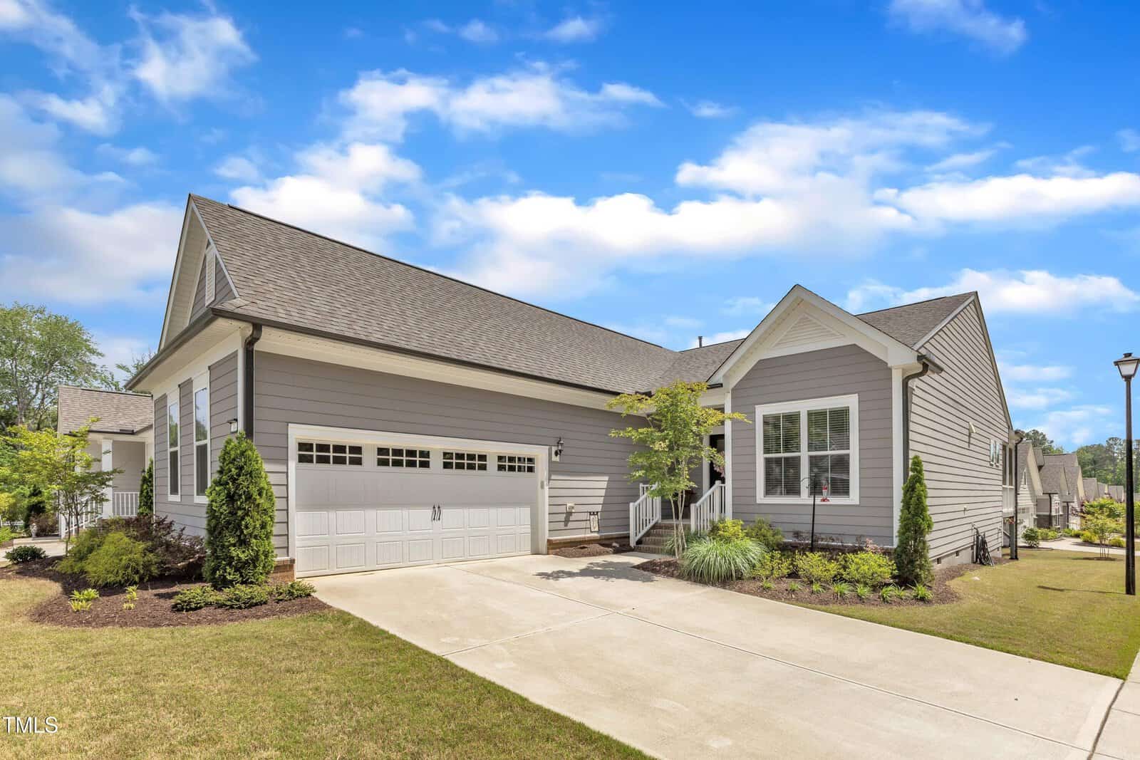 A modern single-story house with gray siding, white trim, and a double garage. The front yard is landscaped with green shrubs and small trees. The sky is blue with scattered clouds.