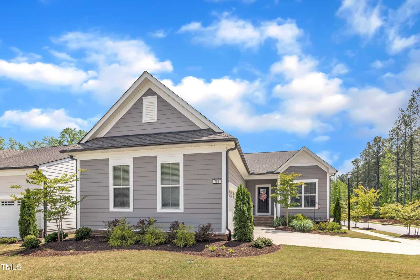 Single-story gray house with white trim, front door under a small porch, manicured lawn, young trees, and driveway. Sky is bright with scattered clouds and tall trees are visible in the background.