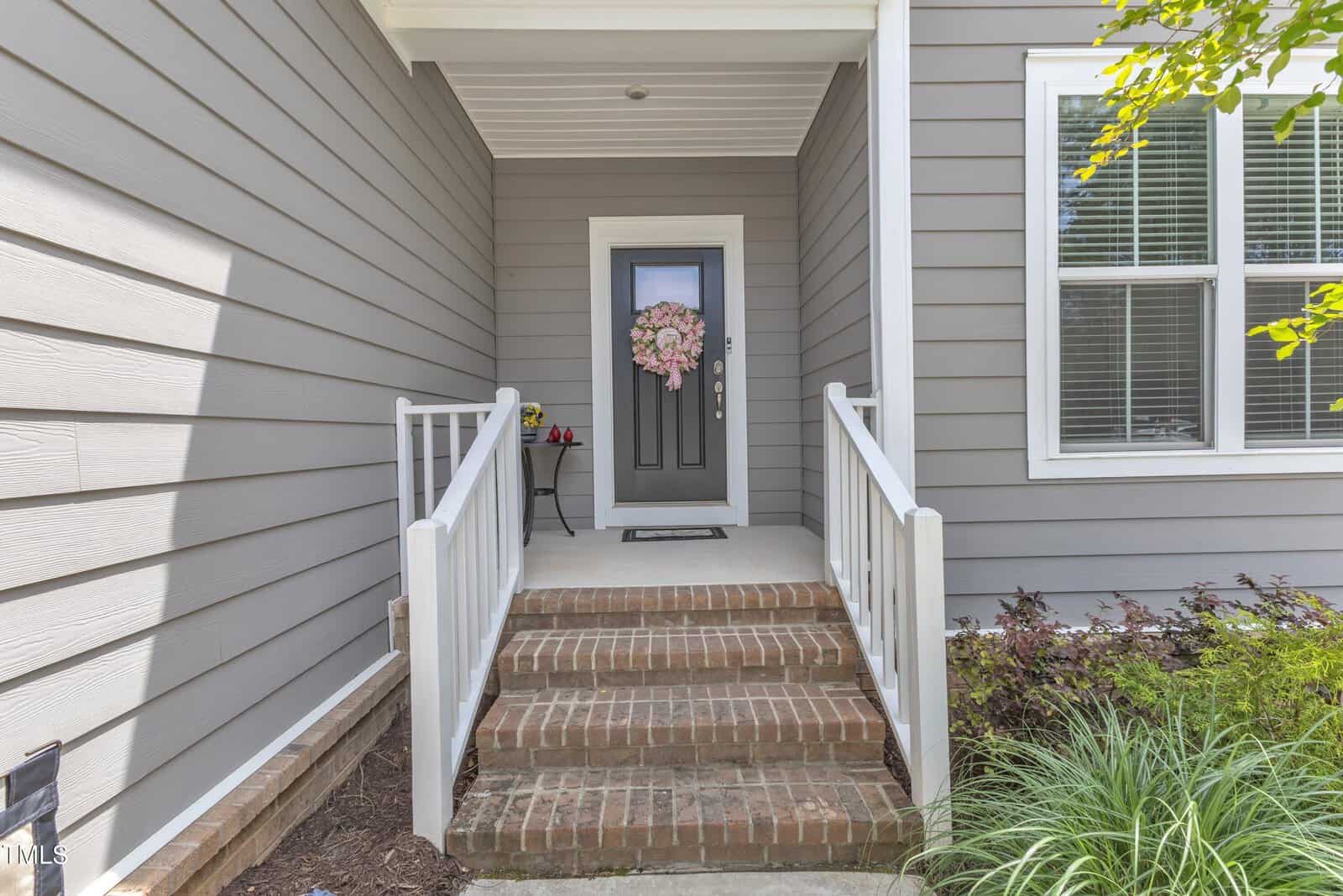 A front porch with gray siding, brick steps, white railings, and a black door decorated with a pink flower wreath. A small table with decor is beside the door. Bushes and plants line the walkway.