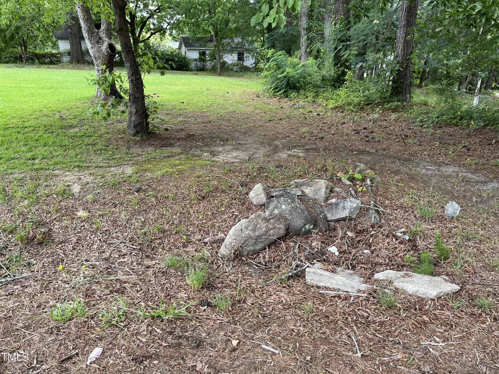 A patch of ground with scattered rocks and sparse grass, bordered by trees and bushes. A grassy yard and a house are visible in the background under a partly cloudy sky.