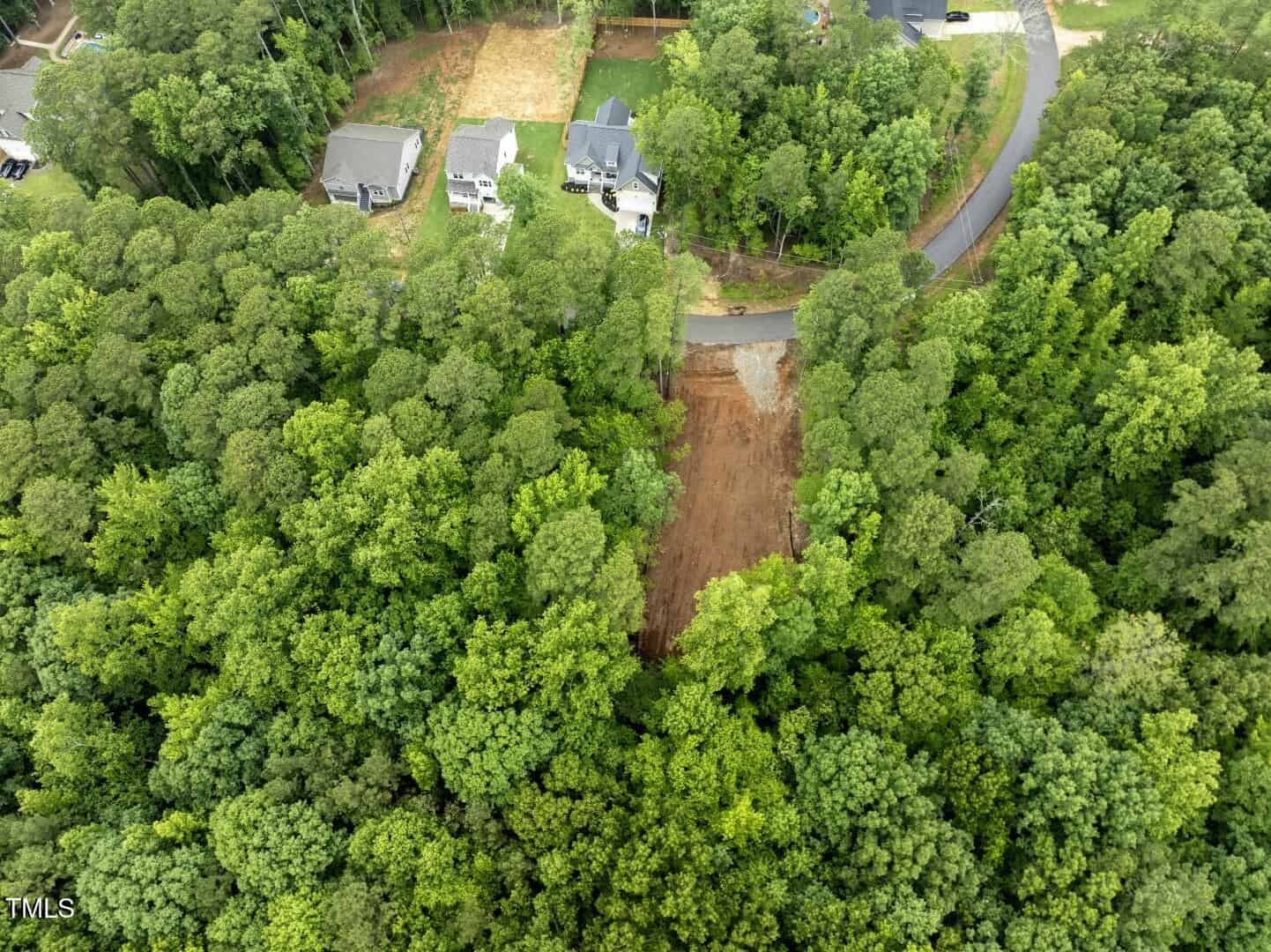 Aerial view of a forested area with a cleared rectangular plot of land, bordered by trees. Nearby are a few houses along a curved road at the top of the image.