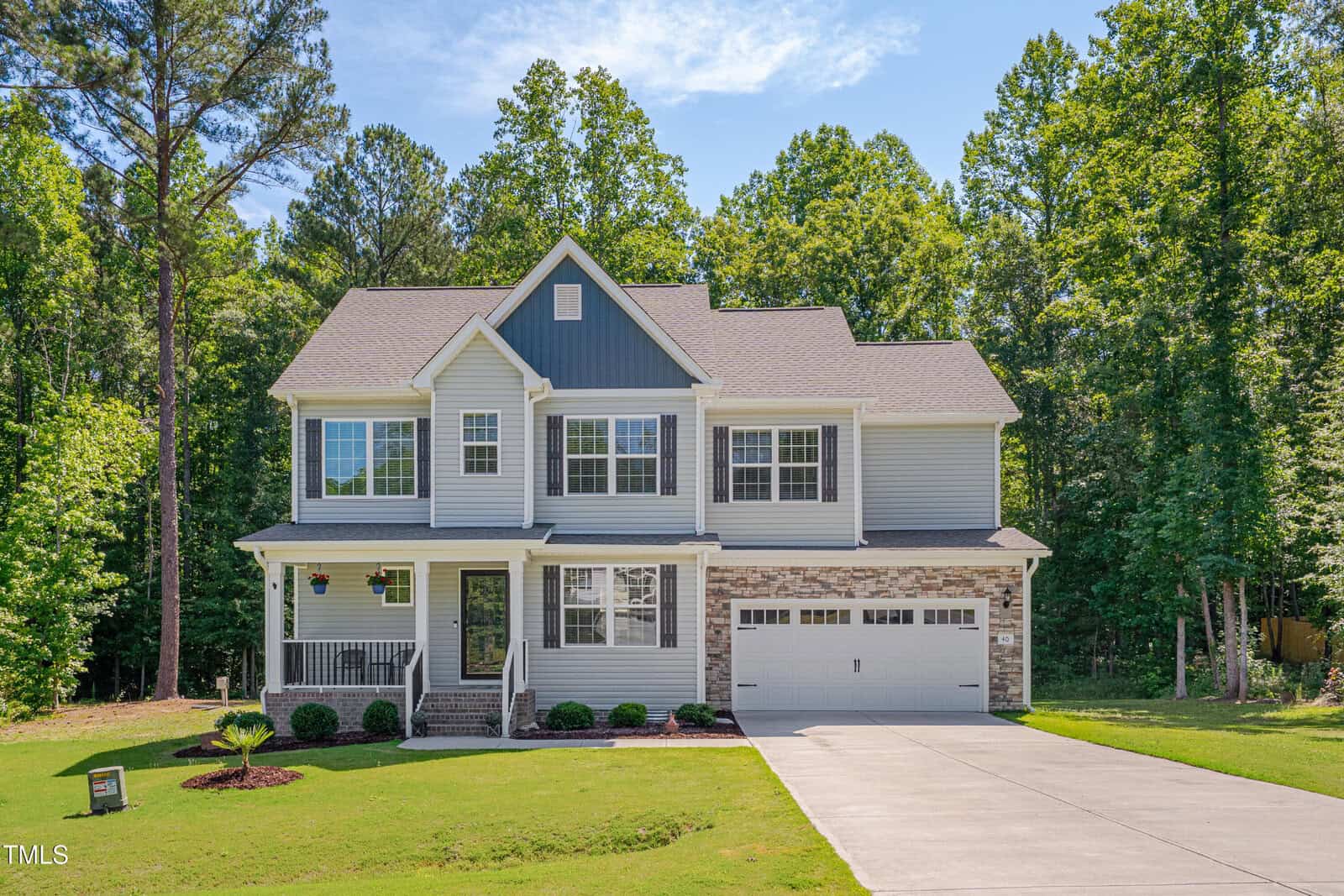 Two-story suburban house with a mix of gray siding and stone accents, attached two-car garage, front porch, and neatly landscaped lawn, surrounded by tall trees under a sunny blue sky.