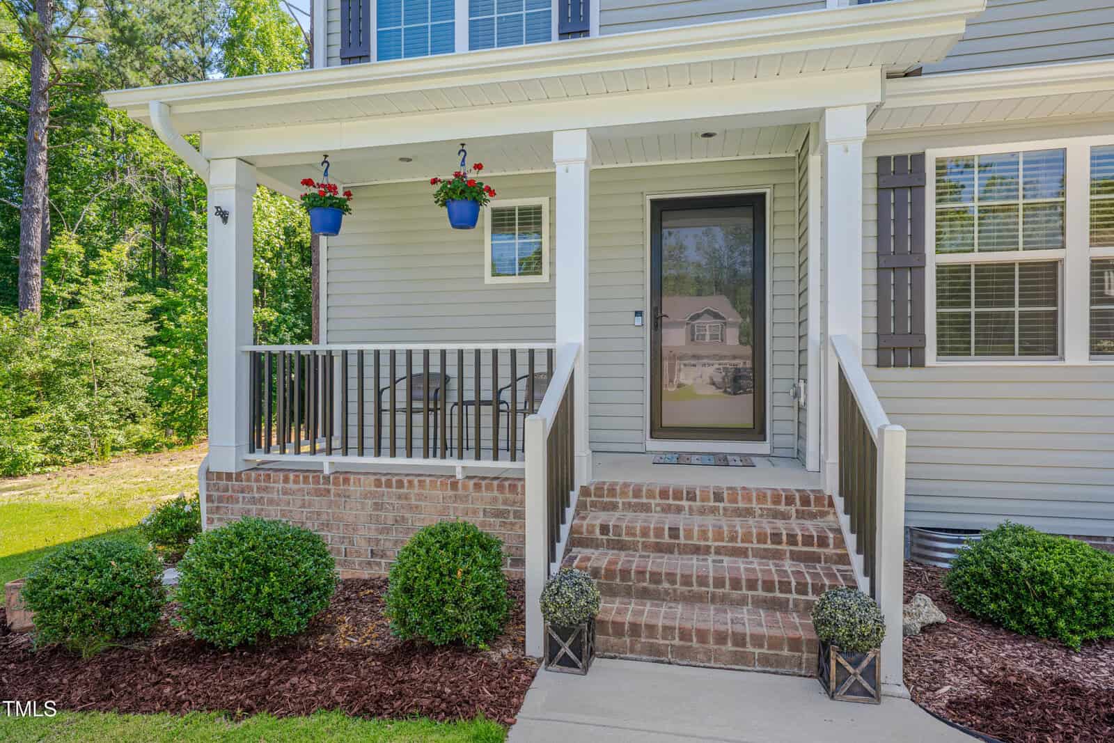 A small front porch with brick steps, a black railing, a glass door, two hanging blue planters with flowers, and a seating area with two chairs. Bushes and mulch line the walkway leading to the porch.