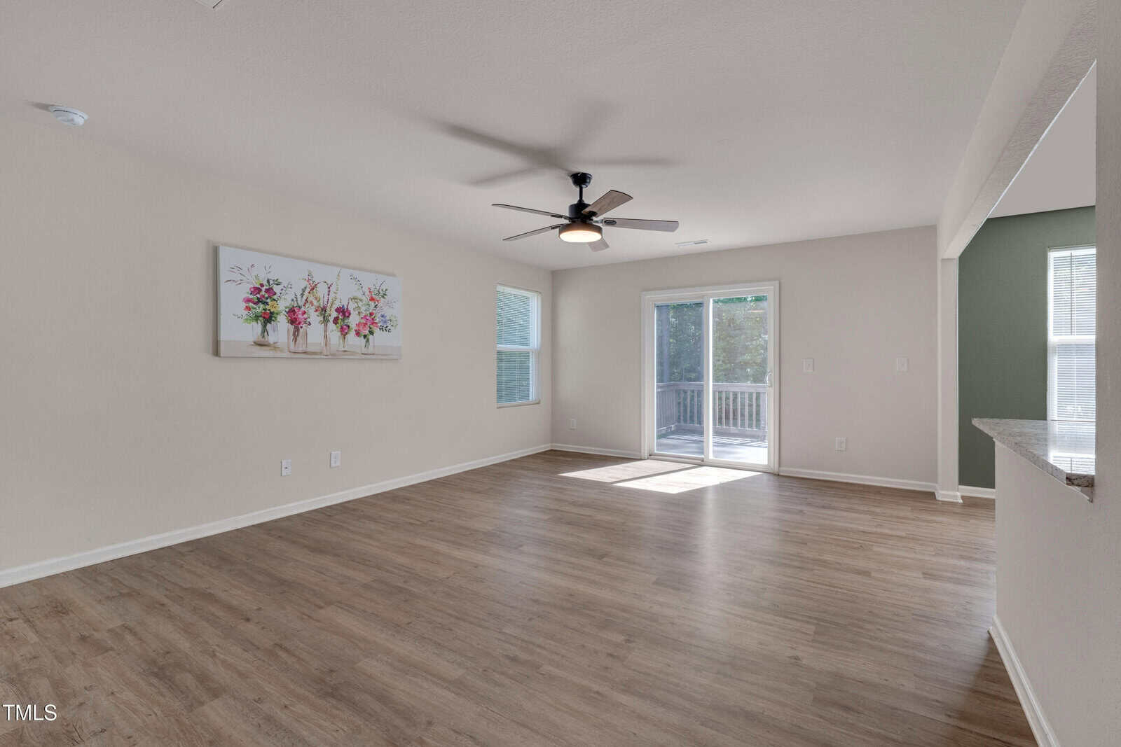 A spacious, empty living room with light-colored walls, wood flooring, a ceiling fan with light, large windows, a glass door leading to a deck, and a floral painting on the wall.