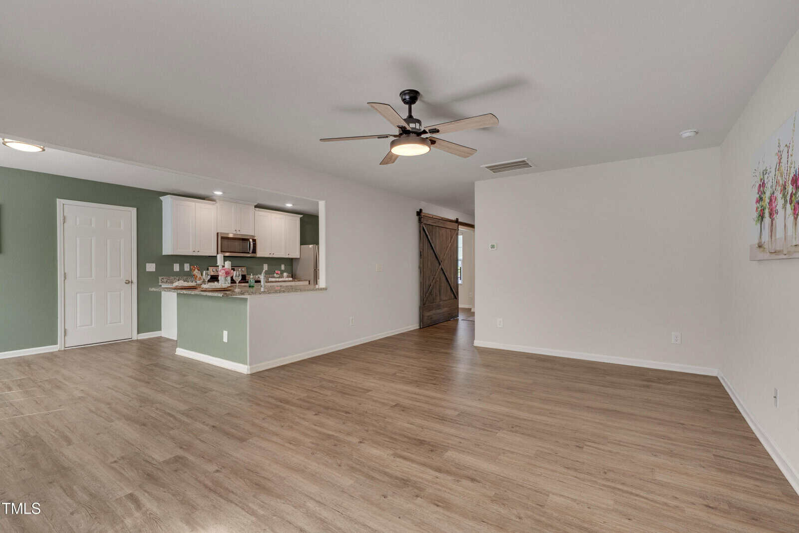 Open-plan living area with light wood flooring, ceiling fan, sage green accent wall, white kitchen cabinets, stainless steel appliances, pantry door, and a sliding barn door. Walls are light beige with a floral artwork visible.