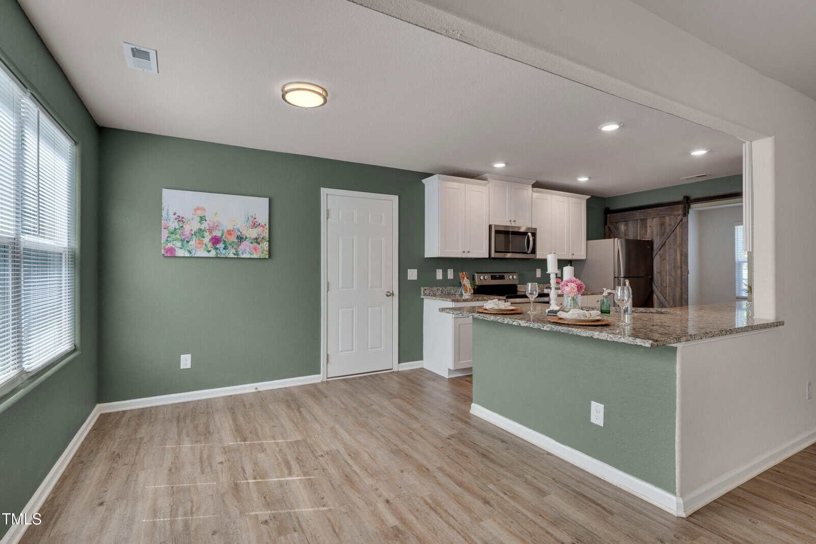 Open kitchen and dining area with green accent wall, wood flooring, white cabinets, granite countertops, stainless steel appliances, a floral painting, and a window allowing natural light into the room.