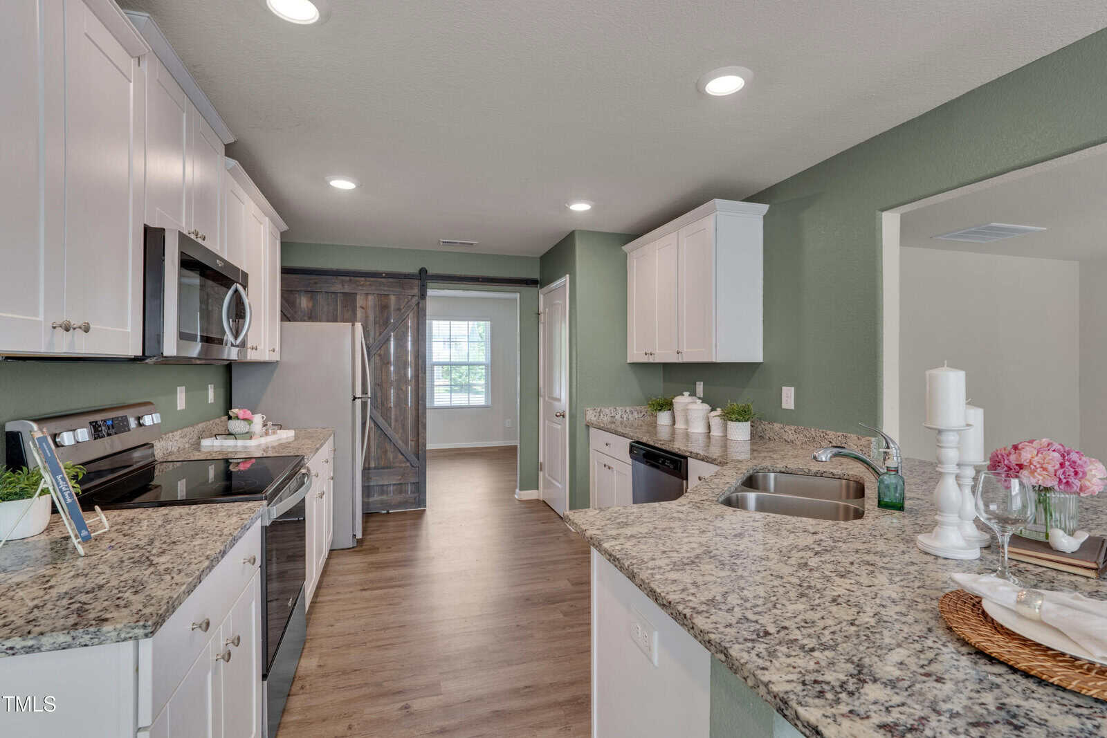 Modern kitchen with white cabinets, granite countertops, stainless steel appliances, and wood-look flooring. A sliding barn door leads to another room, and a window lets in natural light at the far end. Decorative items are on the counters.