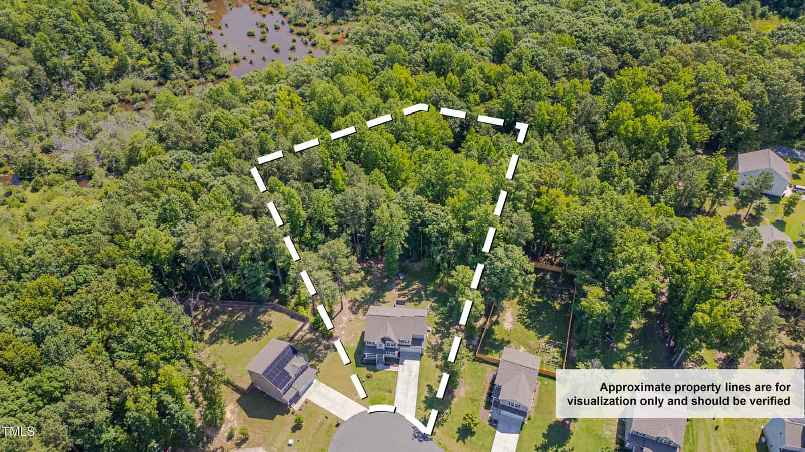 Aerial view of a wooded lot with dashed white lines outlining an approximate triangular property boundary behind a house on a cul-de-sac. Nearby homes and a pond are visible, with a disclaimer in the corner.