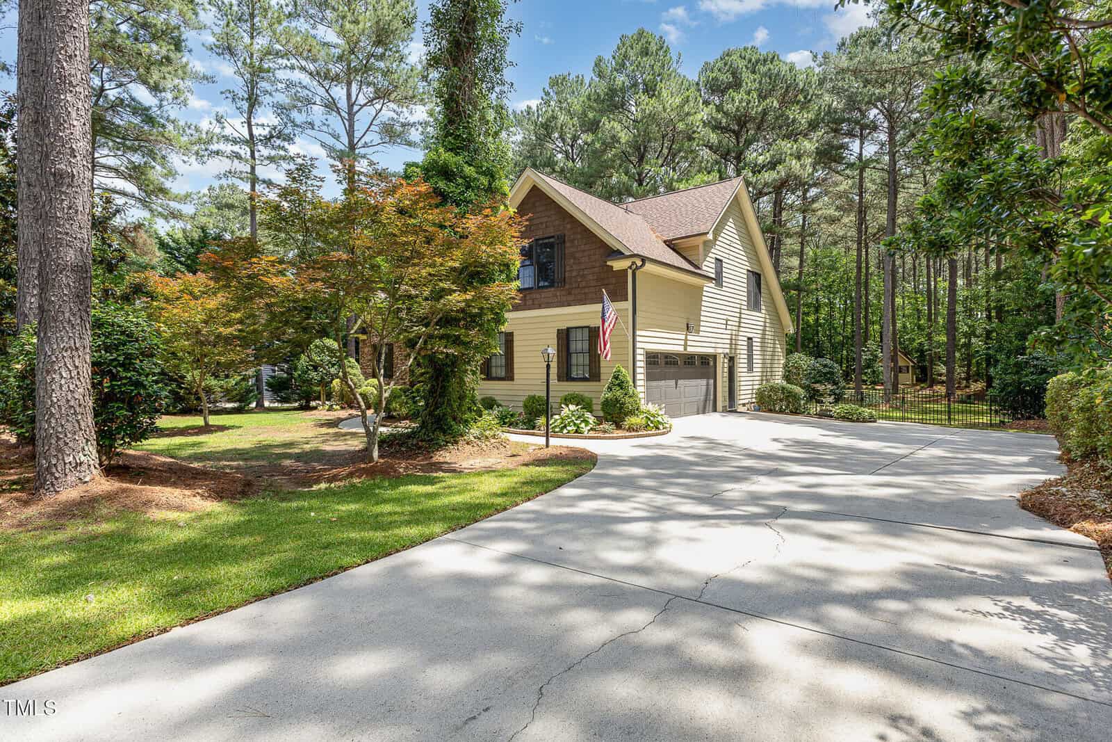 A two-story beige house with a brown roof and attached garage, surrounded by tall trees and green lawn, with a curved driveway and an American flag by the entrance.
