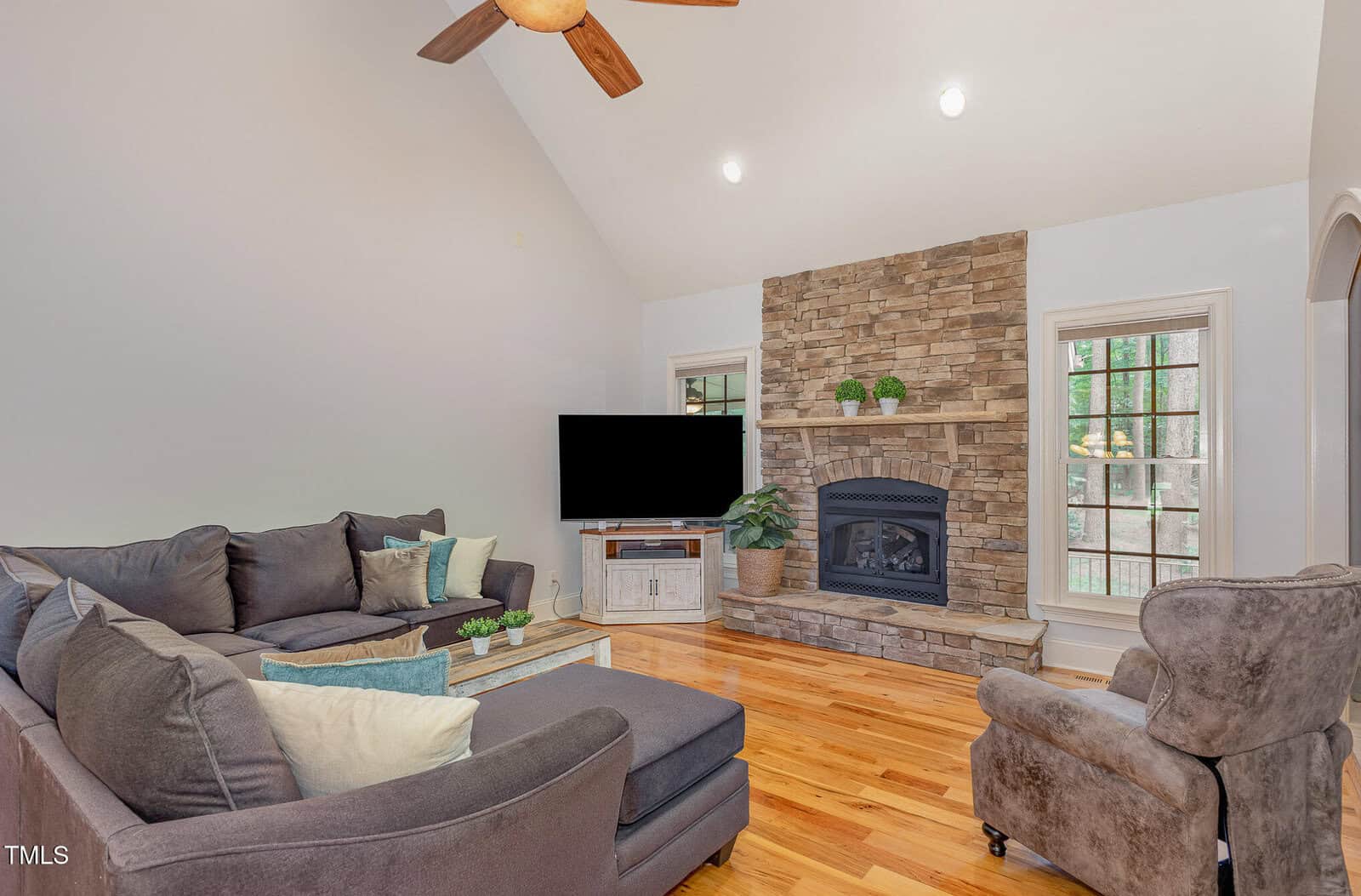A living room with hardwood floors, gray sectional sofa, gray armchair, a coffee table, a mounted TV, and a stone fireplace flanked by windows. There are potted plants on the fireplace mantel and TV stand.