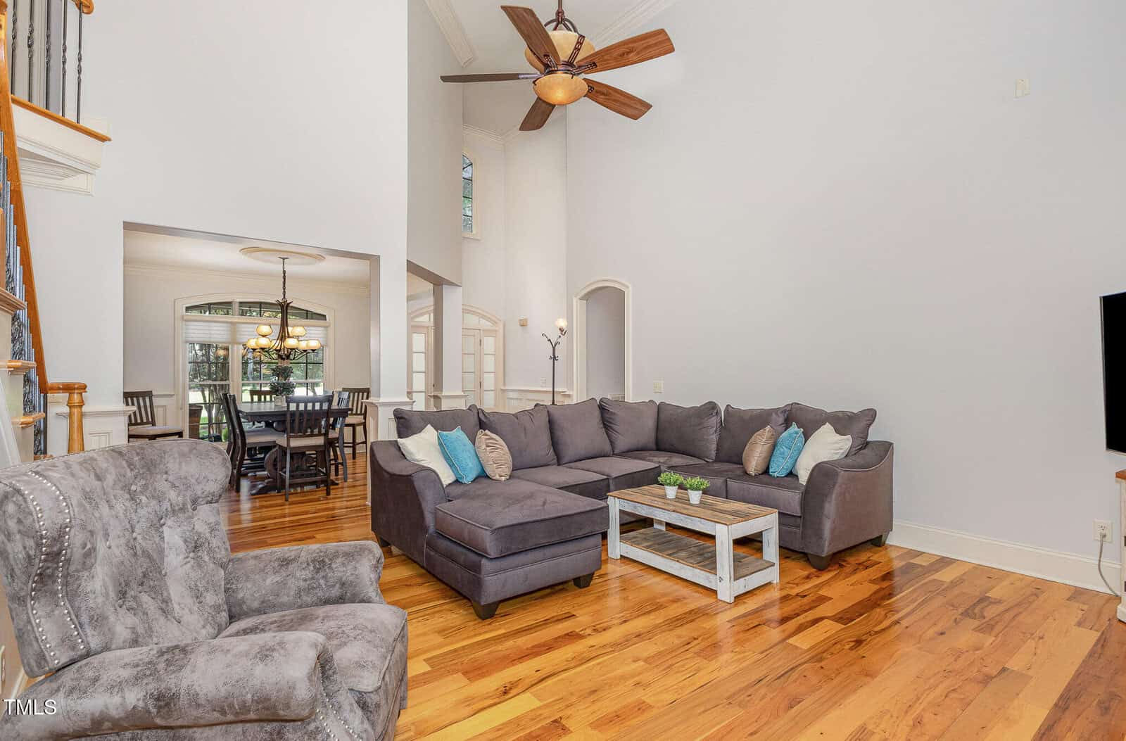 A living room with two gray upholstered sectionals, a matching armchair, a wooden coffee table, hardwood floors, high vaulted ceiling, ceiling fan, and an open view into the dining area.