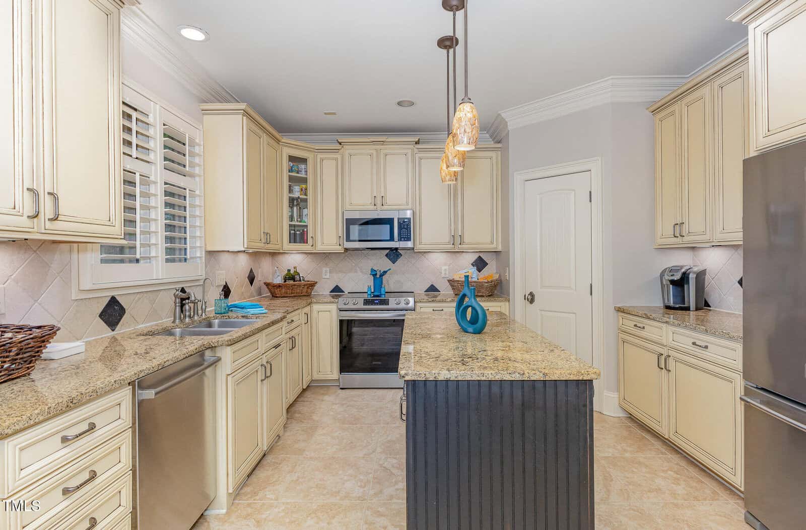 Modern kitchen with cream cabinets, granite countertops, stainless steel appliances, a center island with pendant lights, a window with white shutters, and beige tiled floor. Decorative items and a coffee maker are visible.
