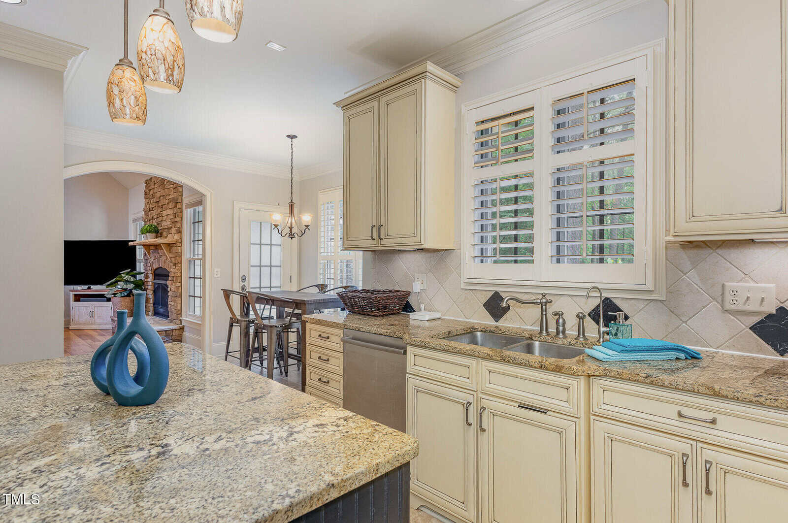 A kitchen with cream cabinets, granite countertops, a double sink, stainless steel appliances, pendant lights, and a dining area with a chandelier visible in the background. A living area and TV are also partially visible.