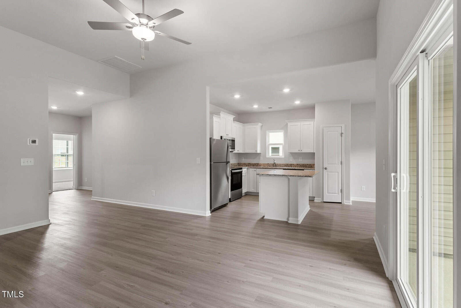 Open-concept living area with light wood flooring, a ceiling fan, and sliding glass doors leading outside. The modern kitchen features white cabinets, stainless steel appliances, and recessed lighting.