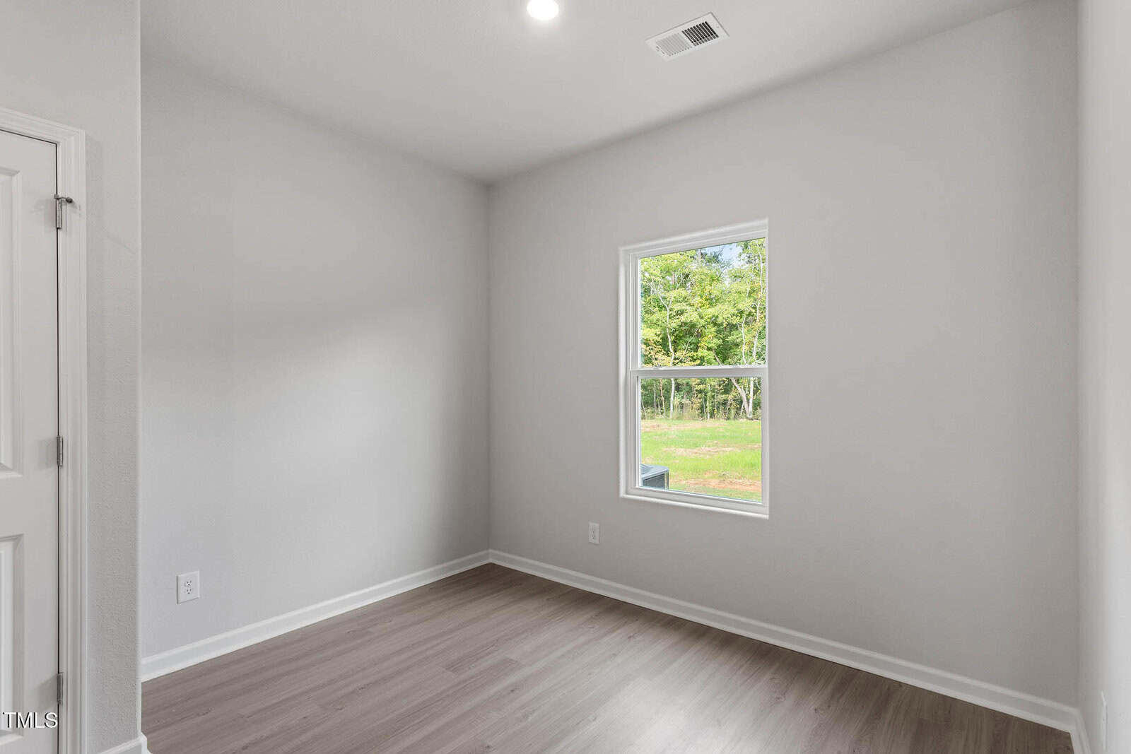 A small, empty room with light gray walls, a white ceiling, wood-style flooring, a single window letting in natural light, and a white door partially visible on the left side.