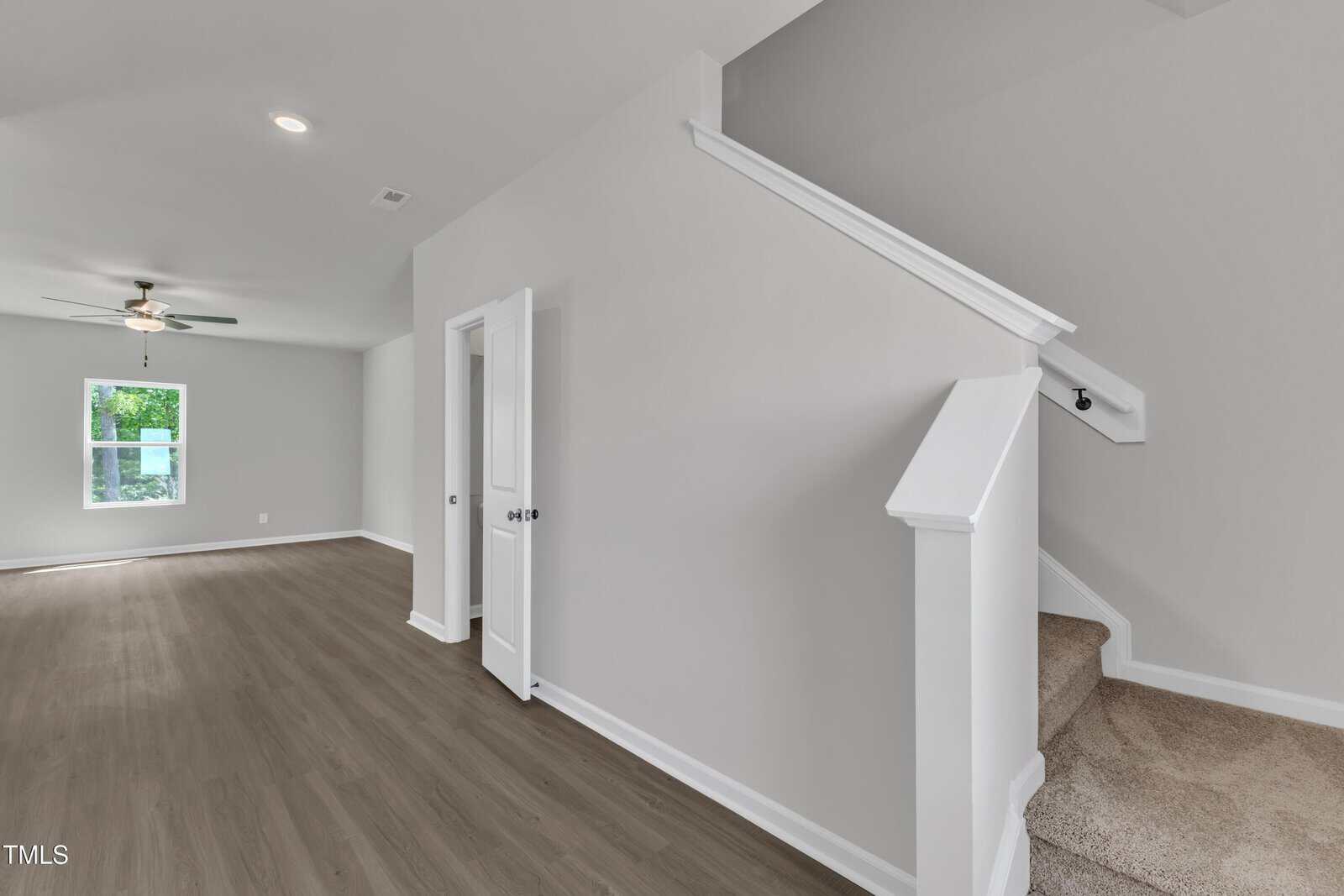 A modern, unfurnished interior shows a room with light gray walls, wood-look flooring, a window, a ceiling fan, and carpeted stairs with a white railing leading to an upper floor. An open door is visible beside the stairs.