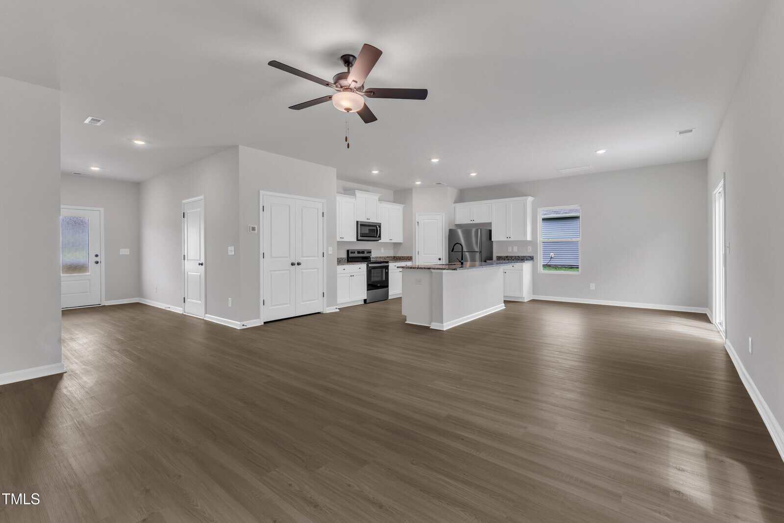 Spacious open-plan kitchen and living area with light gray walls, wood-look flooring, white cabinetry, stainless steel appliances, a ceiling fan, and multiple windows and doors providing natural light.