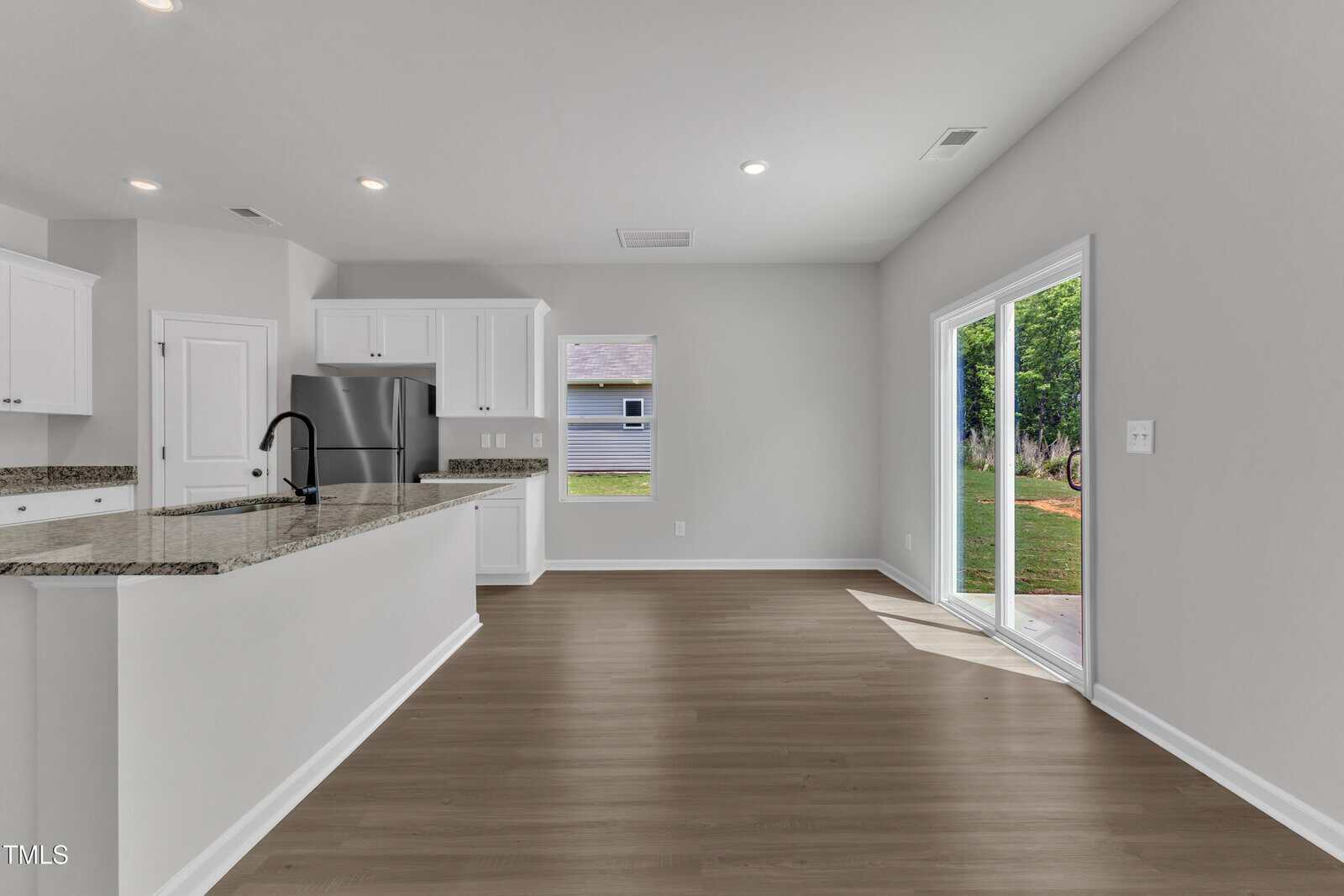 A modern, empty kitchen and dining area with white cabinets, stainless steel appliances, granite countertops, light gray walls, wood-look flooring, a central island, and sliding glass doors leading to a backyard.