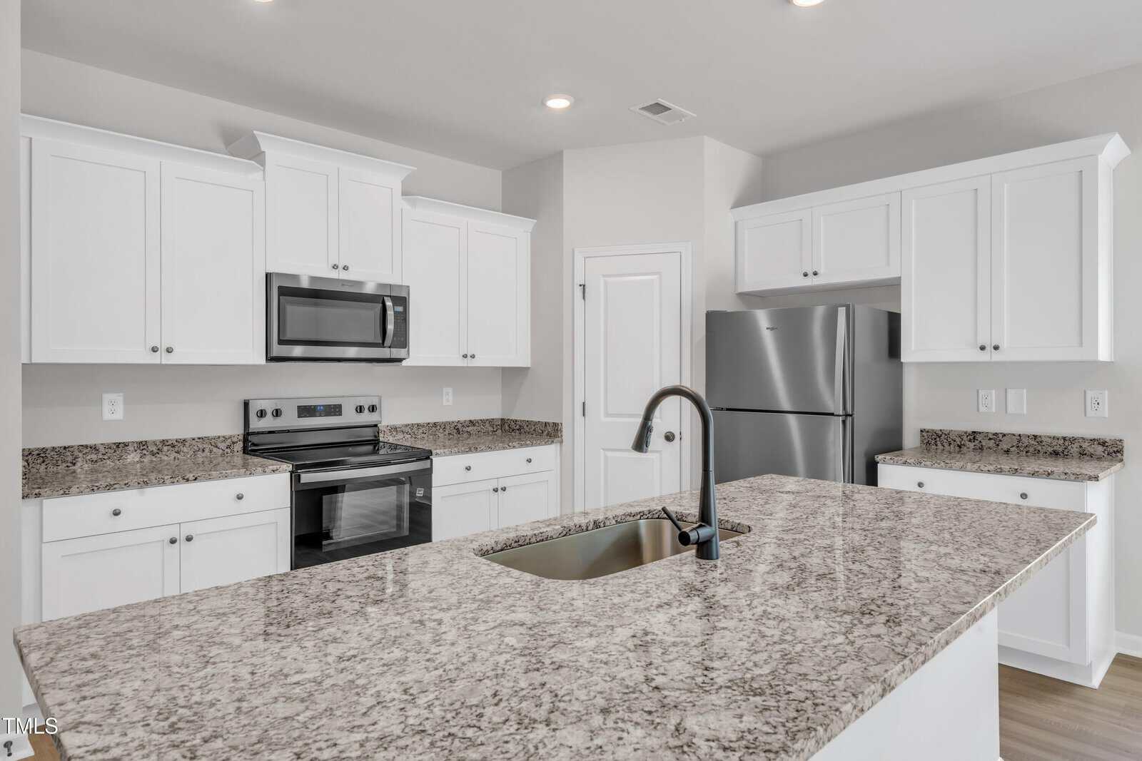 A modern kitchen with white cabinets, stainless steel appliances, granite countertops, an island with a sink, and light gray walls. The space is well-lit with recessed ceiling lights.