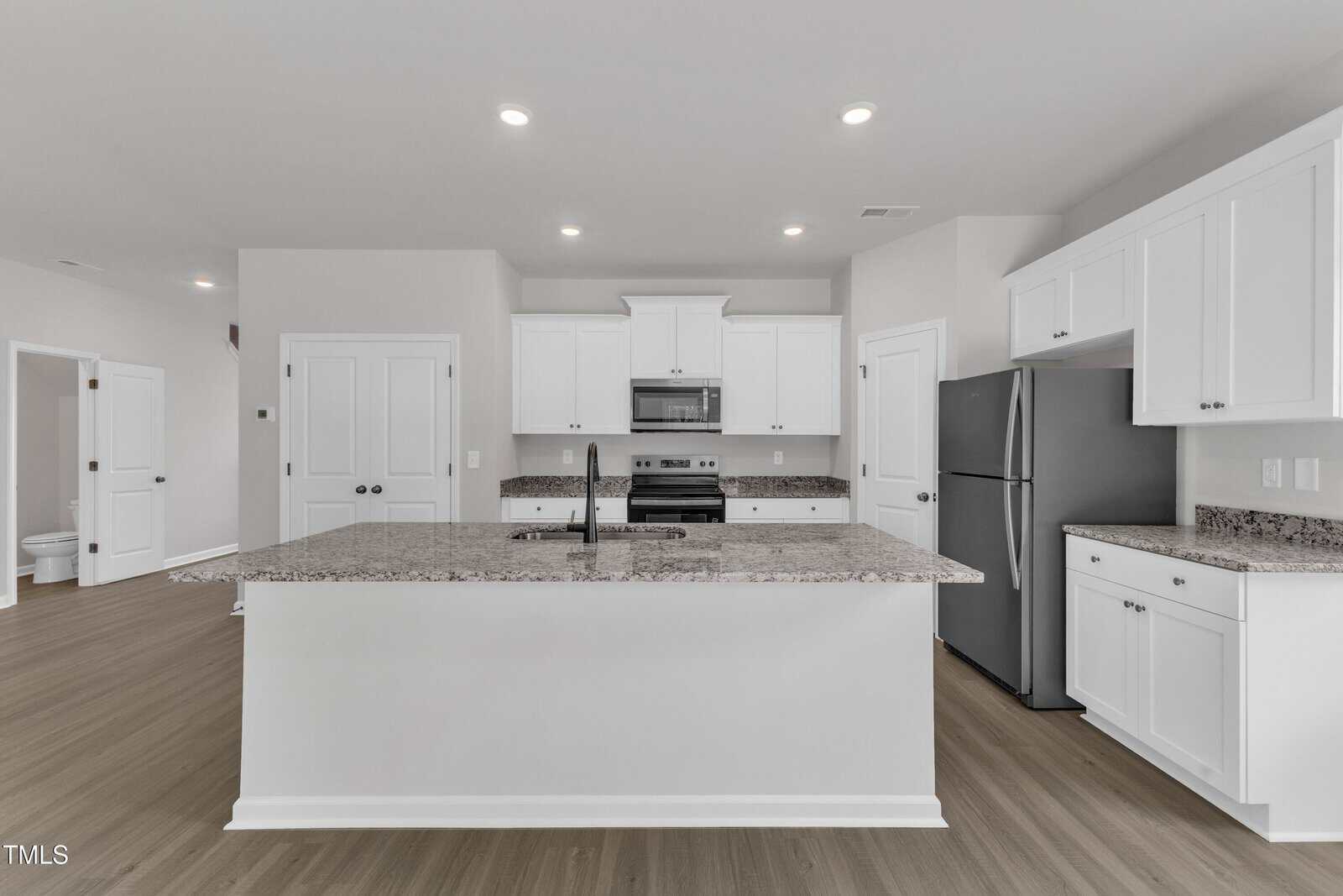 Modern kitchen with white cabinets, granite countertops, stainless steel appliances, a large island with a sink, recessed lighting, and light wood flooring. A doorway and double doors are visible in the background.