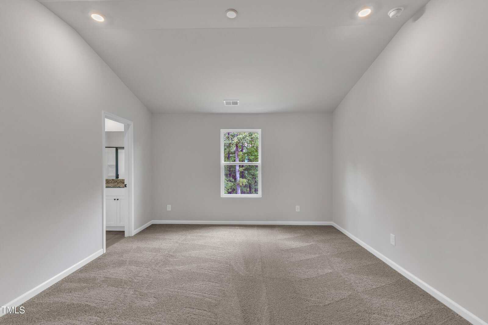 Empty room with light gray walls, beige carpet, white trim, a single window centered on the far wall, ceiling lights, and an open doorway on the left leading to a bathroom with visible countertop and sink.