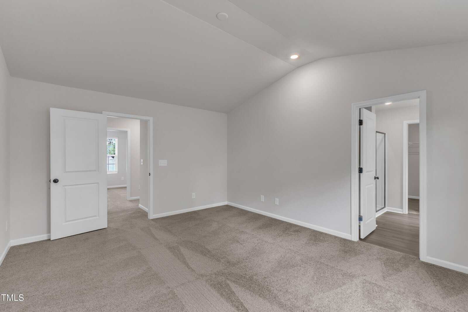 A spacious, empty room with light gray walls, beige carpet, and a vaulted ceiling. Two open doors lead to other rooms, and recessed lighting is visible on the ceiling.