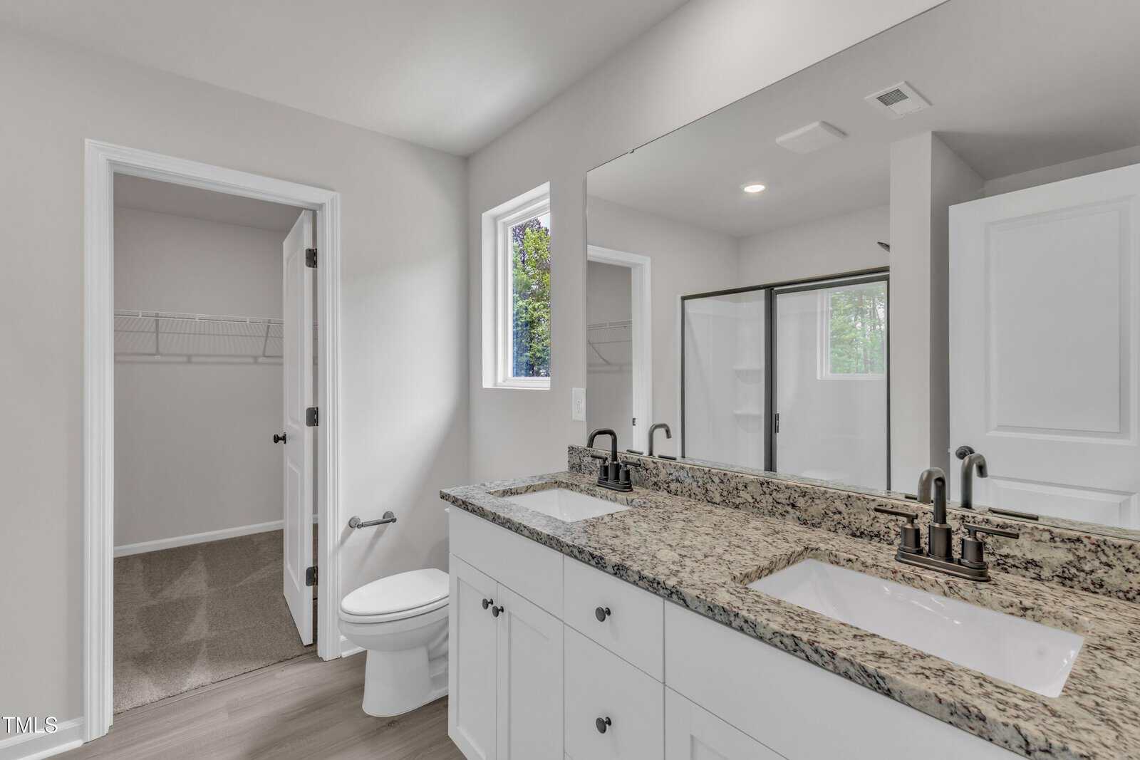 A bathroom with a double-sink vanity, granite countertop, white cabinets, a toilet, a glass-enclosed shower, and a walk-in closet with shelving. Neutral wall colors and natural light from a window.