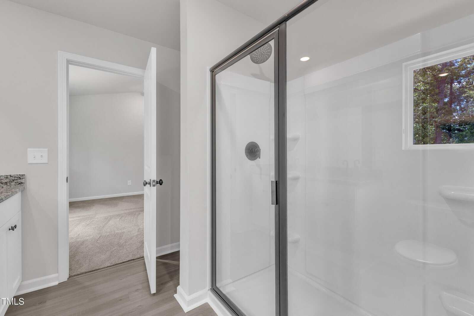 Modern bathroom with a glass shower enclosure, white walls, and a view into a carpeted room through an open door. A small window shows trees outside.