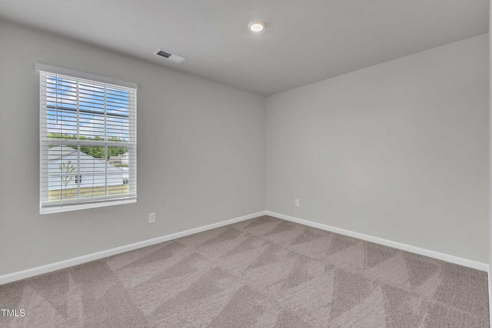 Empty room with light gray walls, a single window with blinds, beige carpet with vacuum lines, and a ceiling light. Natural light is coming in through the window.
