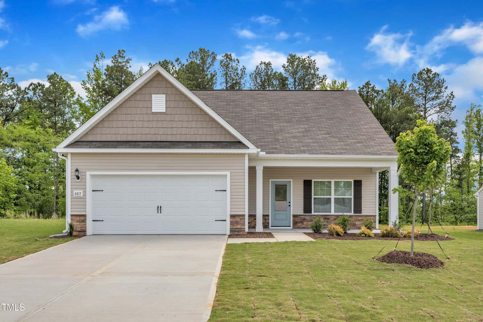 Single-story house with gray siding, a two-car garage, and a small front porch. Located at 503 Benton Drive in Oxford, this home features a neatly manicured lawn, young landscaping, and trees in the background under a bright blue sky with scattered clouds.