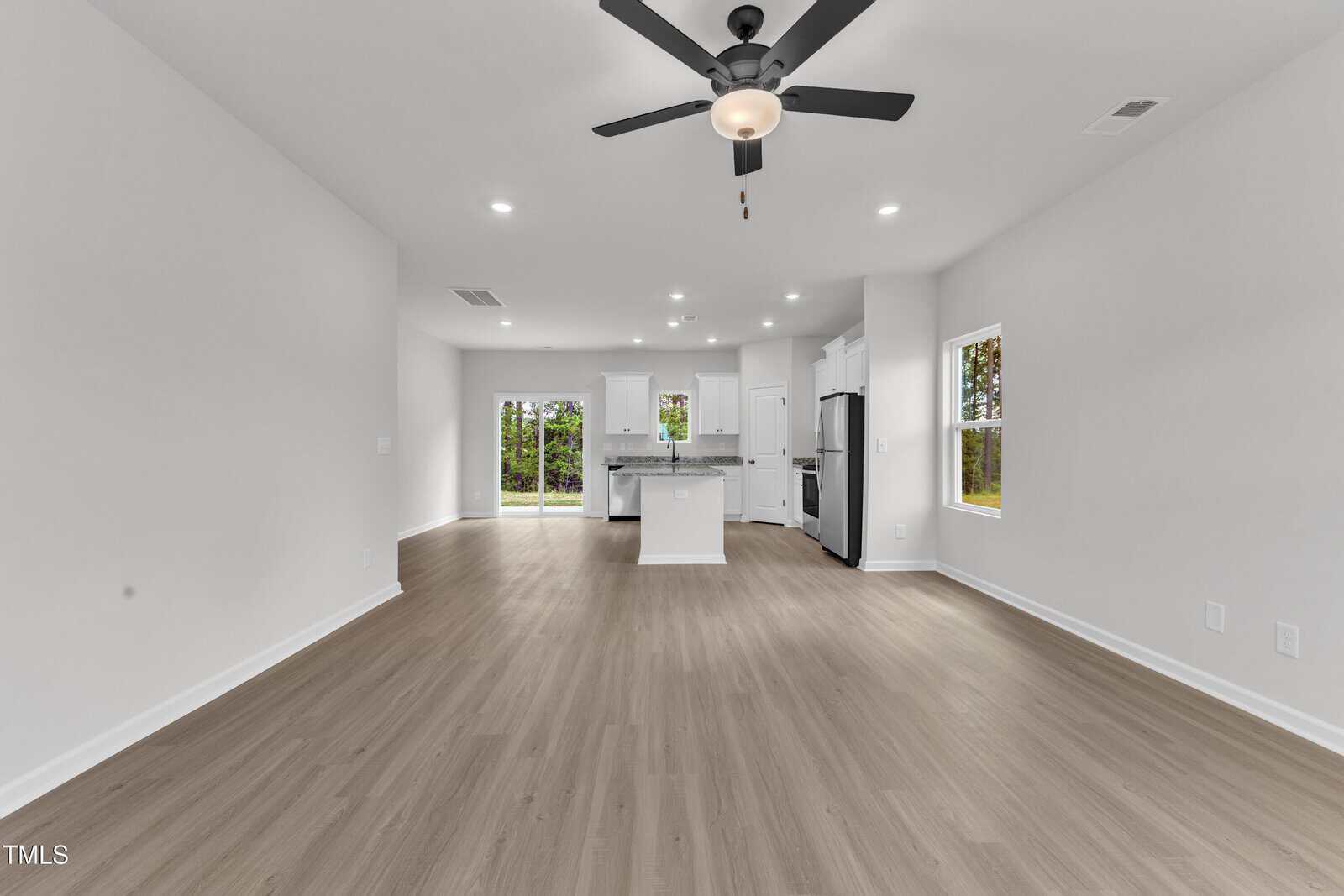 A spacious, empty open-concept living area with light wood floors, white walls, recessed lighting, ceiling fan, and a view of a modern kitchen with an island and stainless steel appliances.