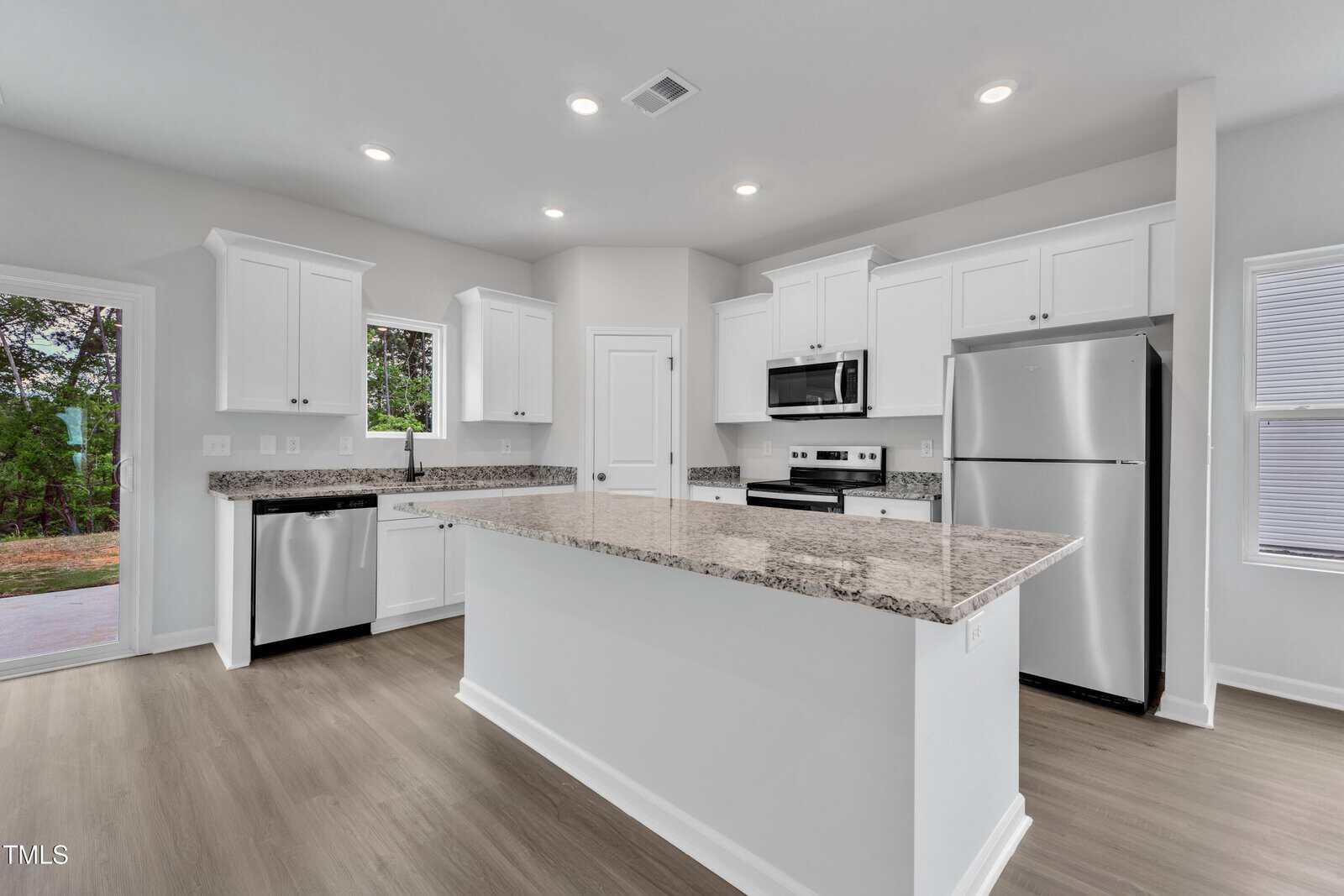 Modern kitchen with white cabinets, stainless steel appliances, a large center island with granite countertop, and light wood flooring. Sliding glass door and windows provide natural light, with a view of greenery outside.