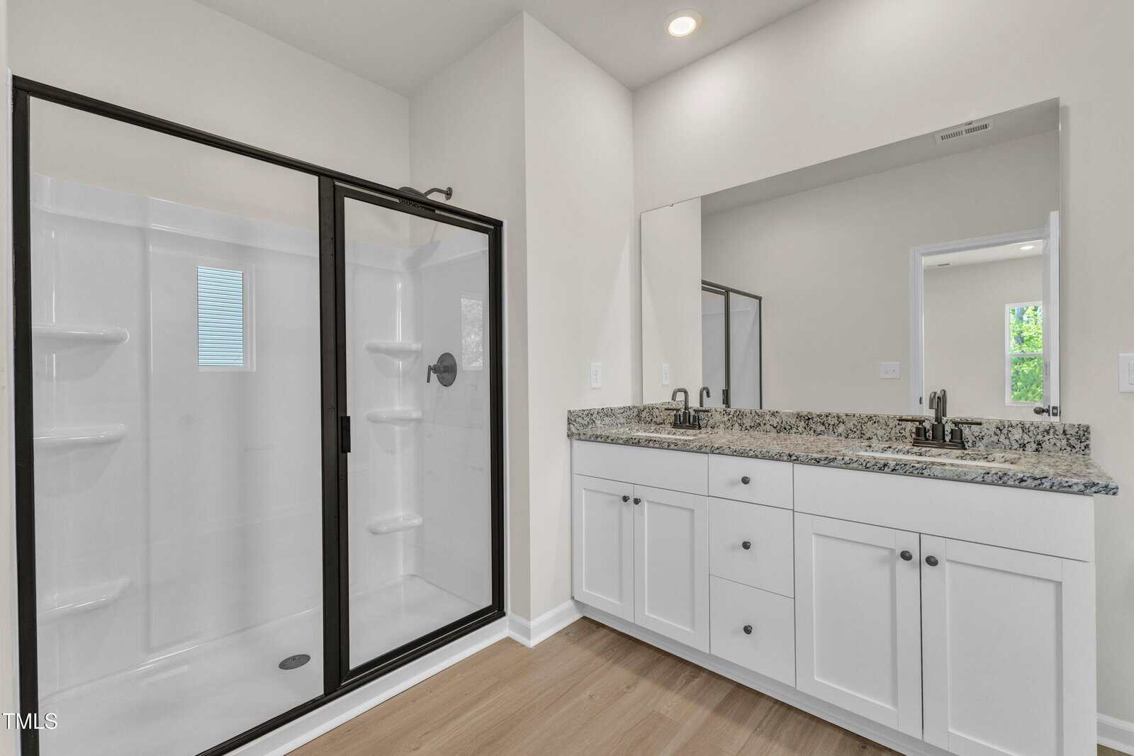 Modern bathroom with a large glass-enclosed shower, white cabinets with granite countertop, dual sinks, and a wide mirror above the vanity. Light wood flooring, neutral walls, and natural light from a window.