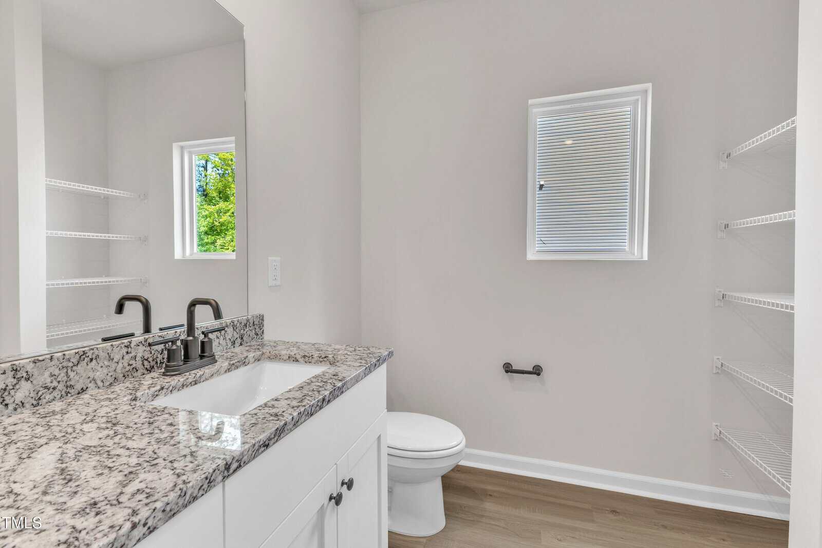 Modern bathroom with a granite countertop sink, white cabinets, wall-mounted black faucet, open shelving, small window with blinds, toilet, and light wood flooring. Walls and trim are painted white.