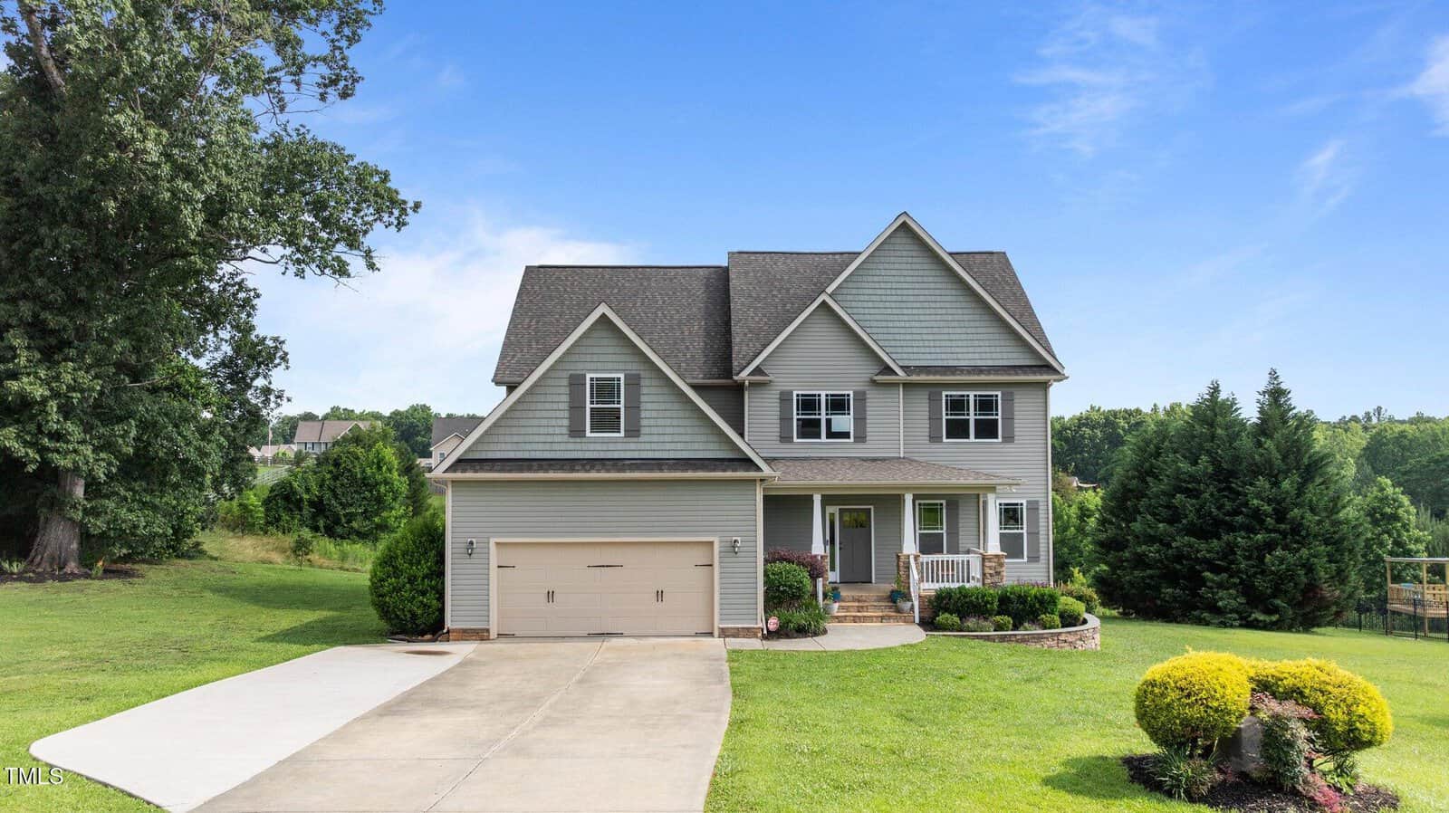 Two-story suburban house with gray siding, a double garage, small front porch, and neatly landscaped yard with green grass and bushes. A concrete driveway leads up to the house. Trees are in the background under a clear blue sky.