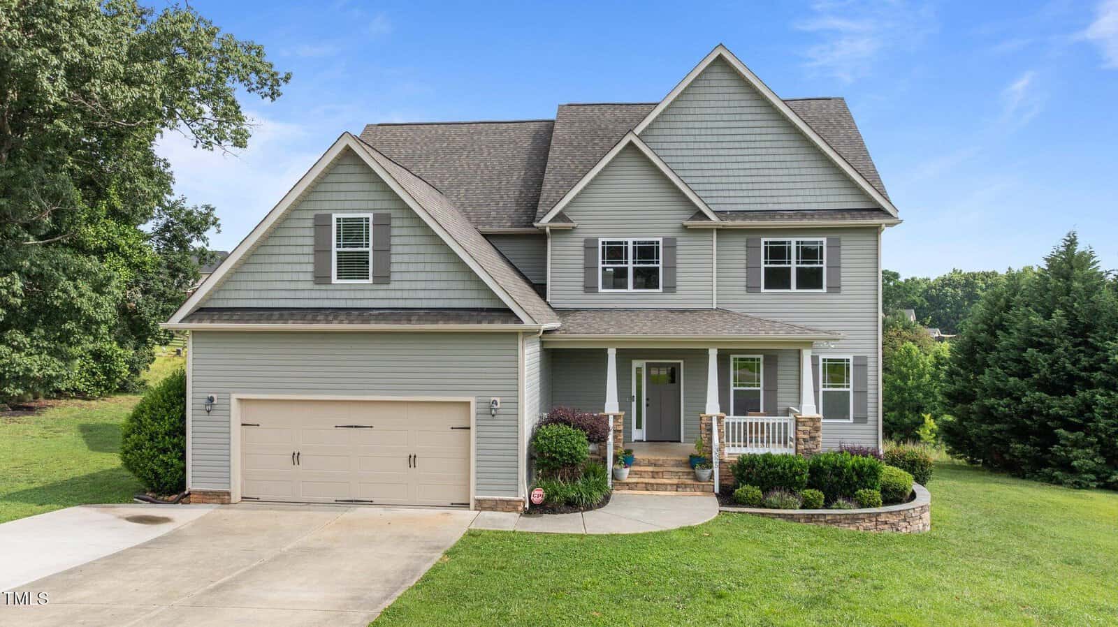 Charming two-story suburban house at 3525 Daisy Lane, Wake Forest, with gray siding, double garage, front porch with columns, small shrubs and neat lawn, all set against a backdrop of trees under a blue sky.