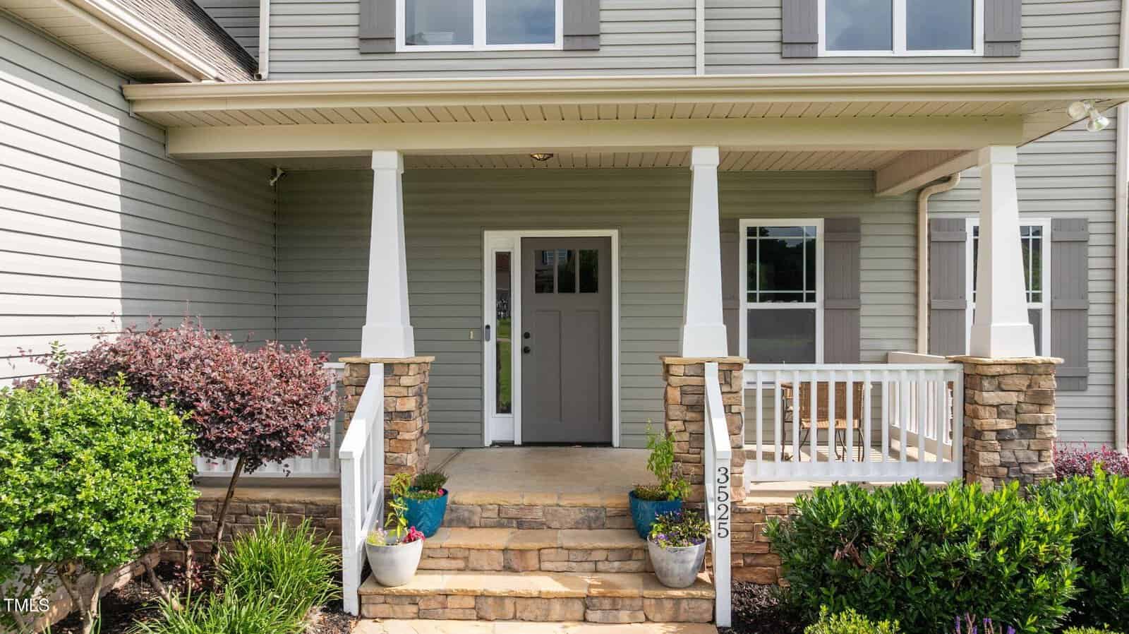 A front porch of a house with stone steps, white pillars, and a gray front door. Potted plants and shrubs line the steps, and the house exterior features gray siding and double windows.