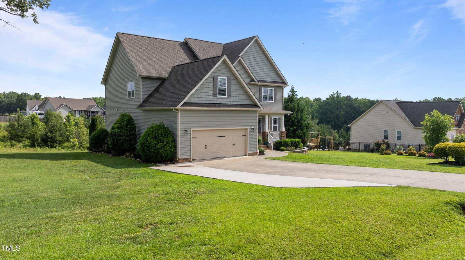 Two-story suburban house with gray siding and an attached two-car garage. The home is set on a large, well-maintained lawn with a concrete driveway, surrounded by other houses and greenery under a blue sky.