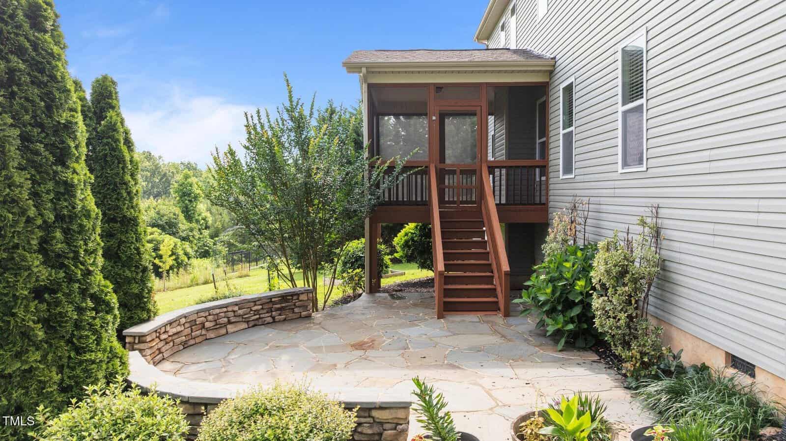 A stone patio with surrounding landscaping is shown next to a house with beige siding. Wooden stairs lead up to a screened porch. Tall shrubs and trees are visible in the background.