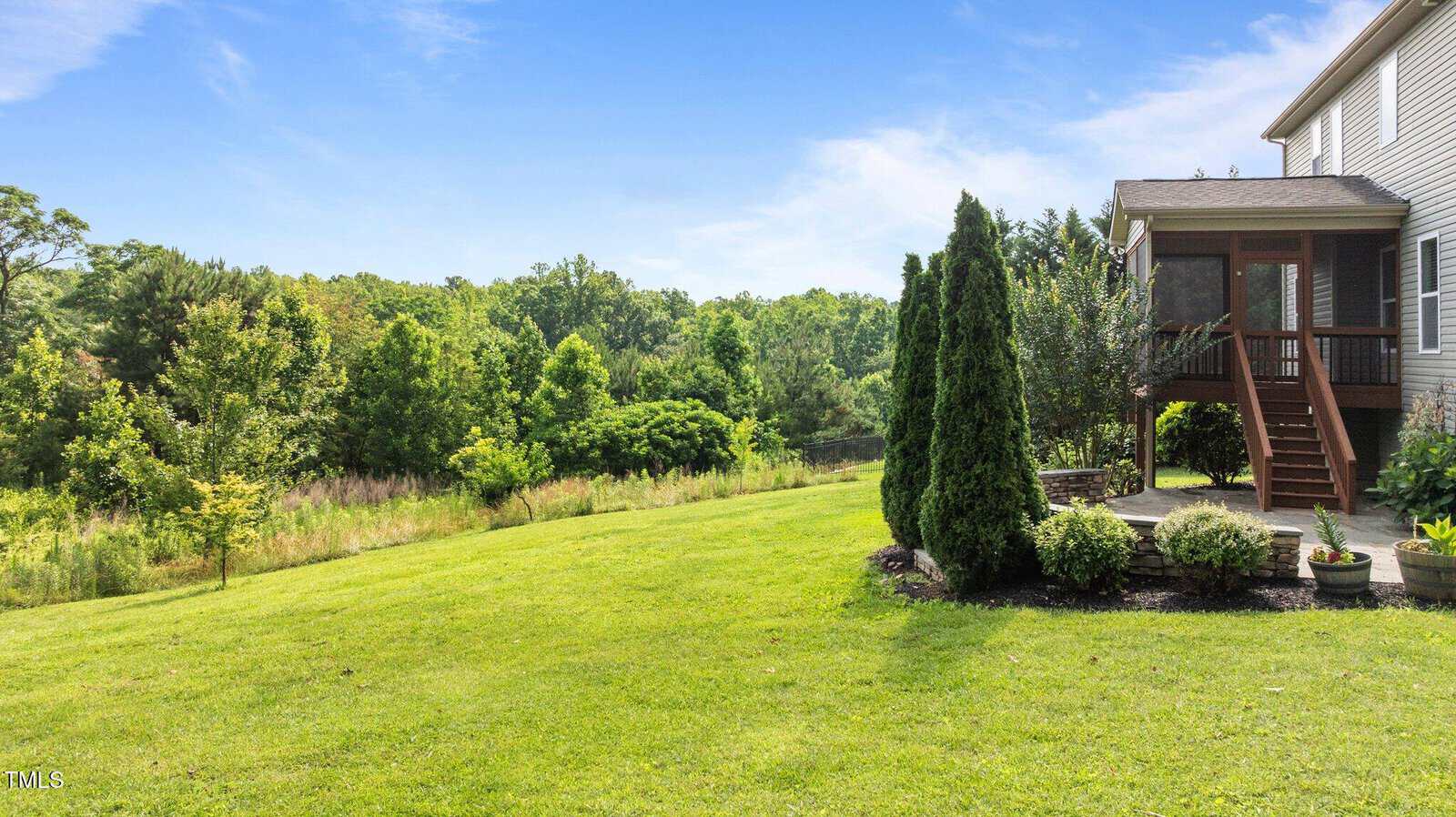 A well-maintained backyard with green grass, trees, and shrubs, adjacent to a two-story house with a screened porch and wooden steps leading down to the yard. Blue sky and wooded area are visible in the background.
