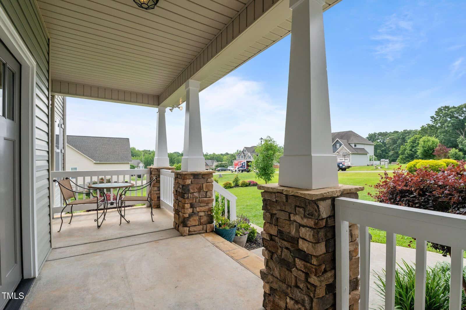 A covered front porch with white railings, stone columns, and a small metal table with two chairs overlooks a suburban neighborhood with green lawns and houses under a clear sky.