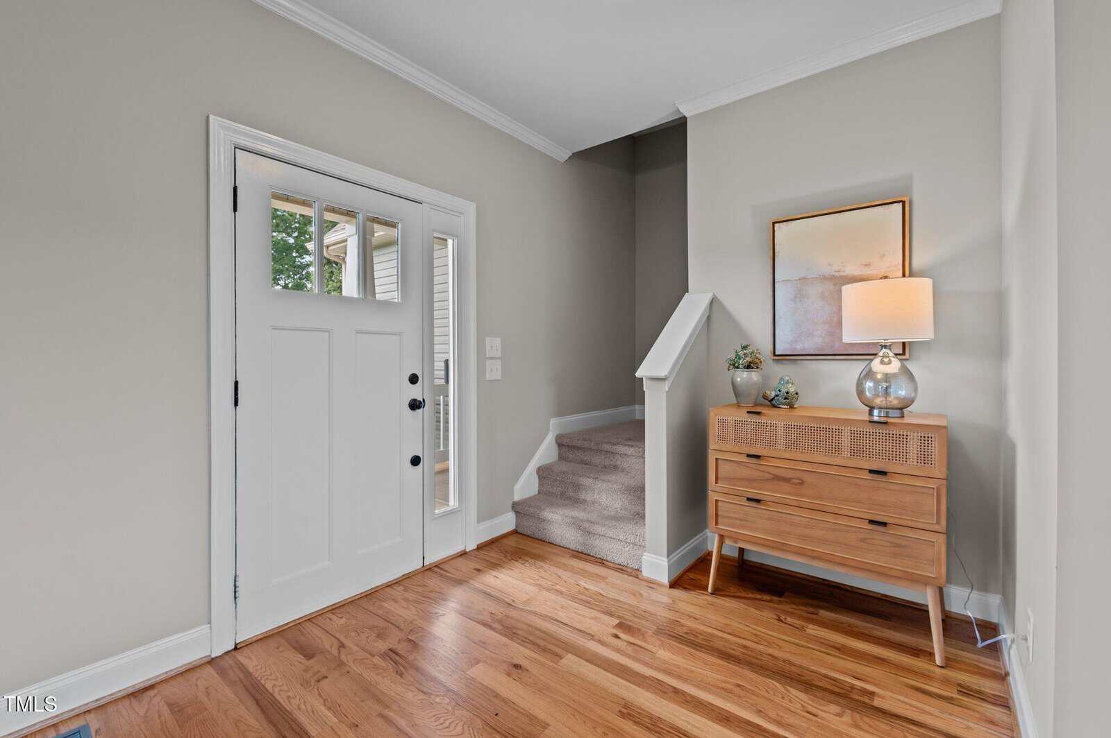 A modern entryway with light hardwood floors, a white front door with glass panels, carpeted stairs, a wooden console table with a lamp, small plants, and a framed artwork on a light gray wall.