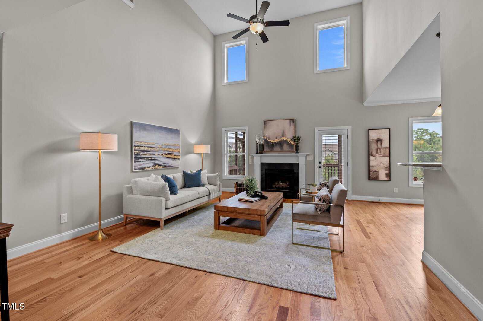 A modern living room with light gray walls, hardwood floors, a white sofa, two armchairs, a wooden coffee table, a fireplace, floor lamp, and large windows allowing natural light into the space.